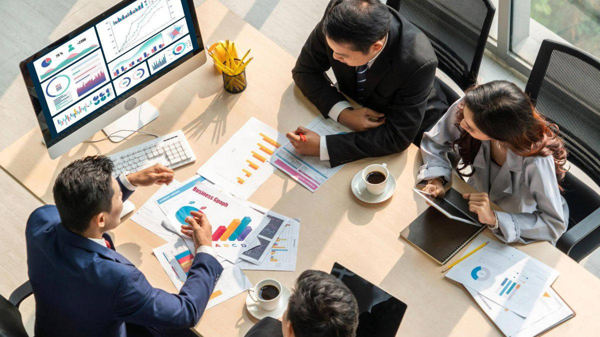 Work 桌布 壁紙 — Overhead view of four business professionals in suits collaborating around a conference table filled with colorful charts and data visualizations.