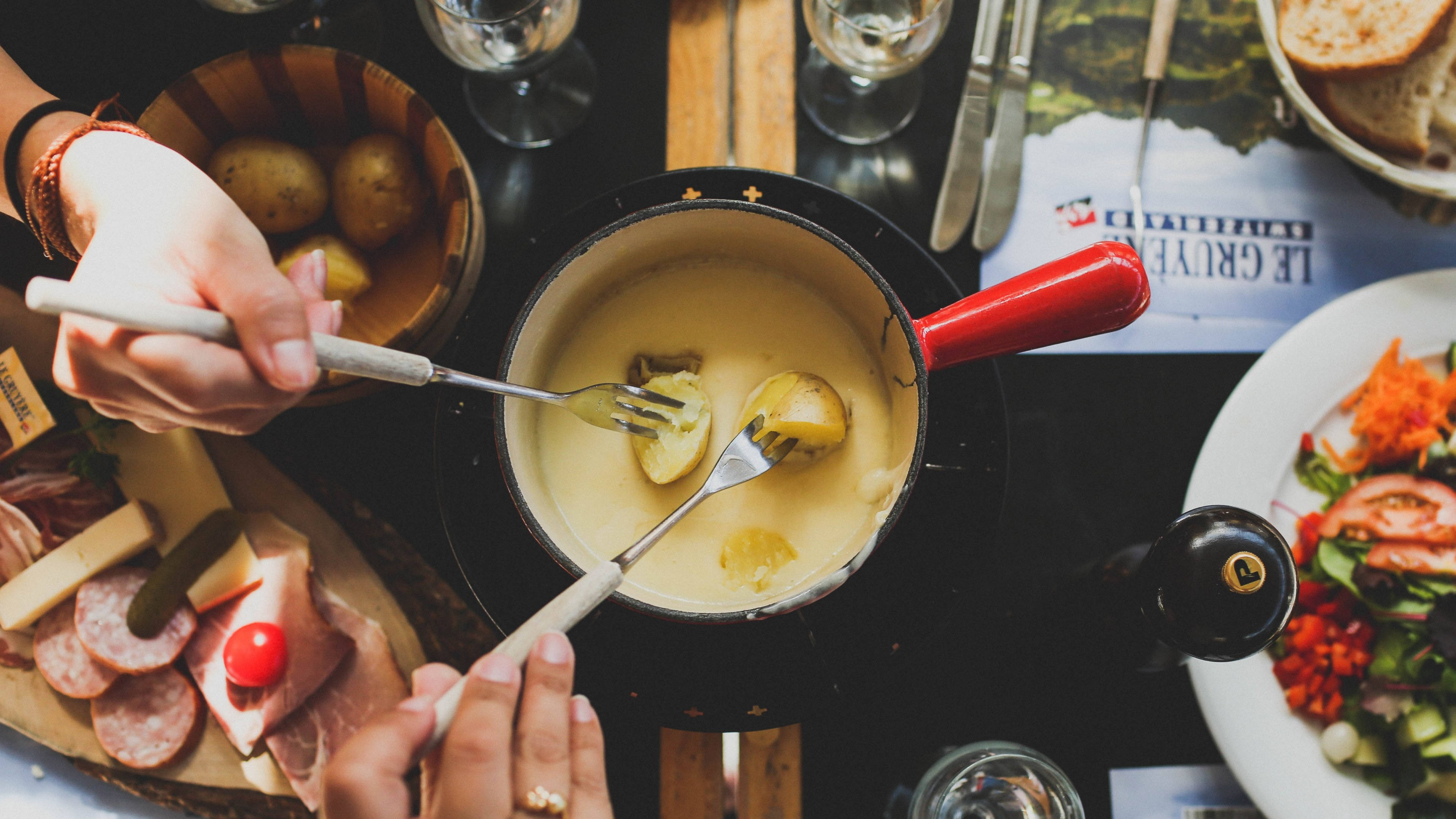 Cheese 桌布 壁紙 — Overhead view of hands dipping boiled potatoes into a bubbling cheese fondue pot surrounded by meats bread and salad.