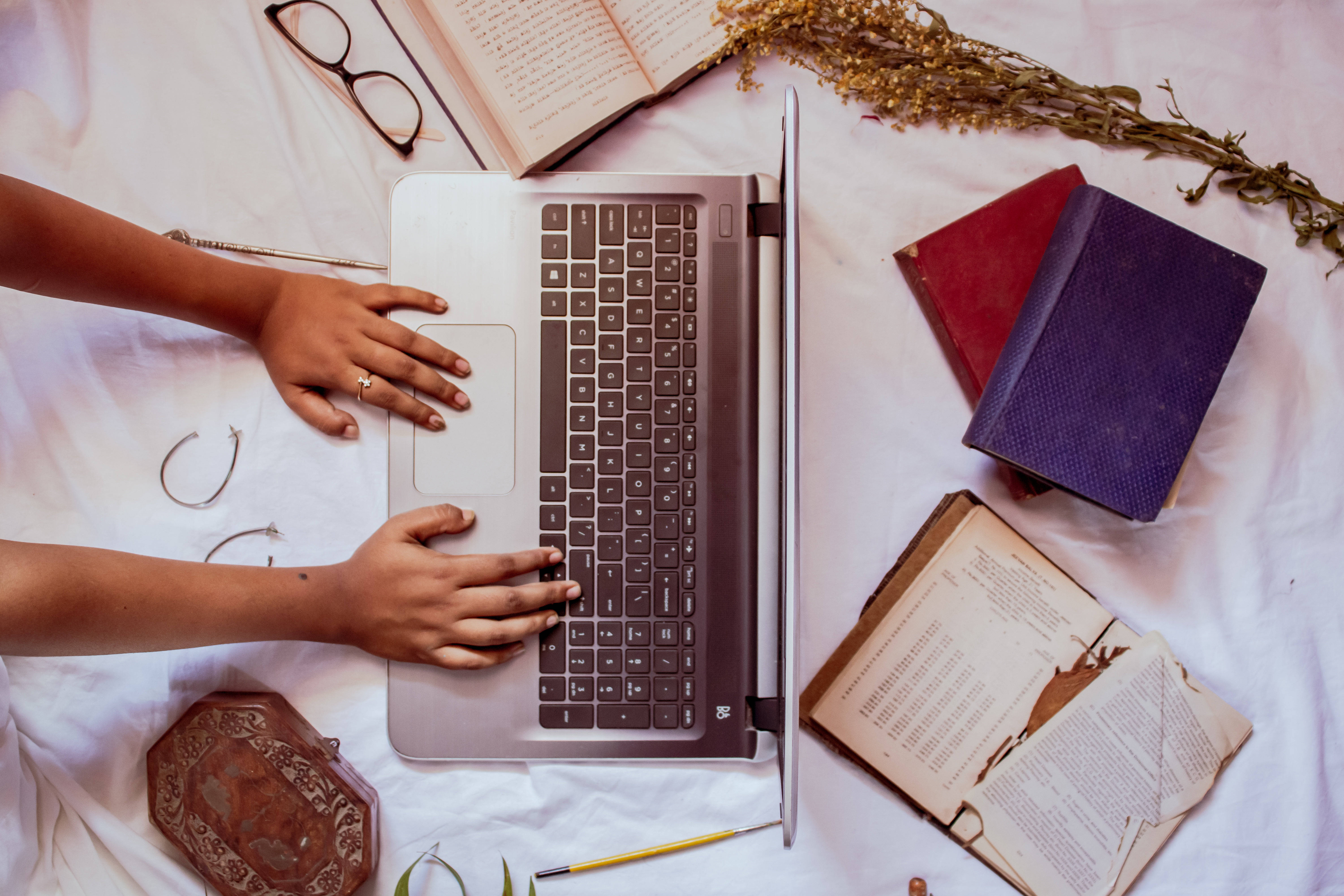 筆記型電腦 桌上型電腦 桌布 壁紙 — Overhead view of hands typing on a laptop surrounded by open books dried flowers and glasses on a white bedspread.