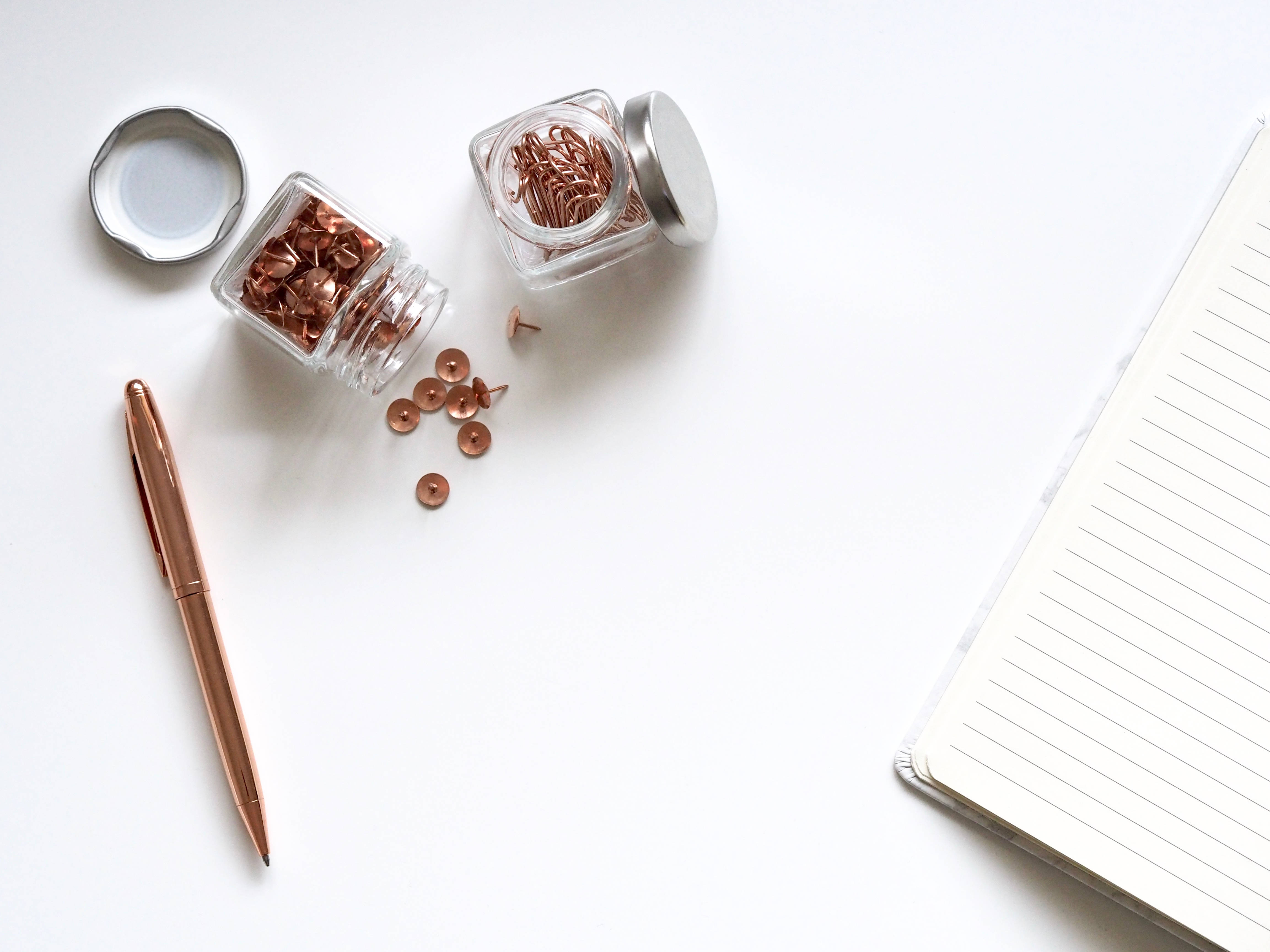 玫瑰金 桌布 壁紙 — Overhead view of rose gold push pins and pen beside a lined notebook on a clean white surface.