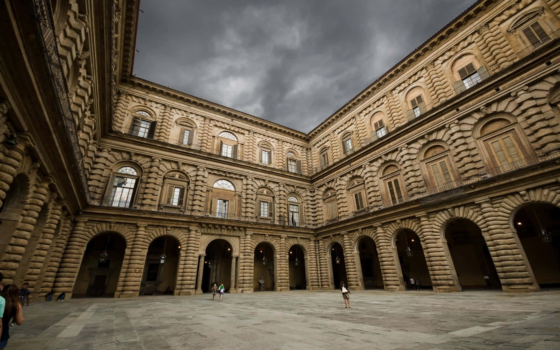 建築 桌布 壁紙 — Palazzo Medici Riccardi 的宏偉石頭庭院，在戲劇性的暴風雲下，有幾個訪客在拱形柱廊下行走。