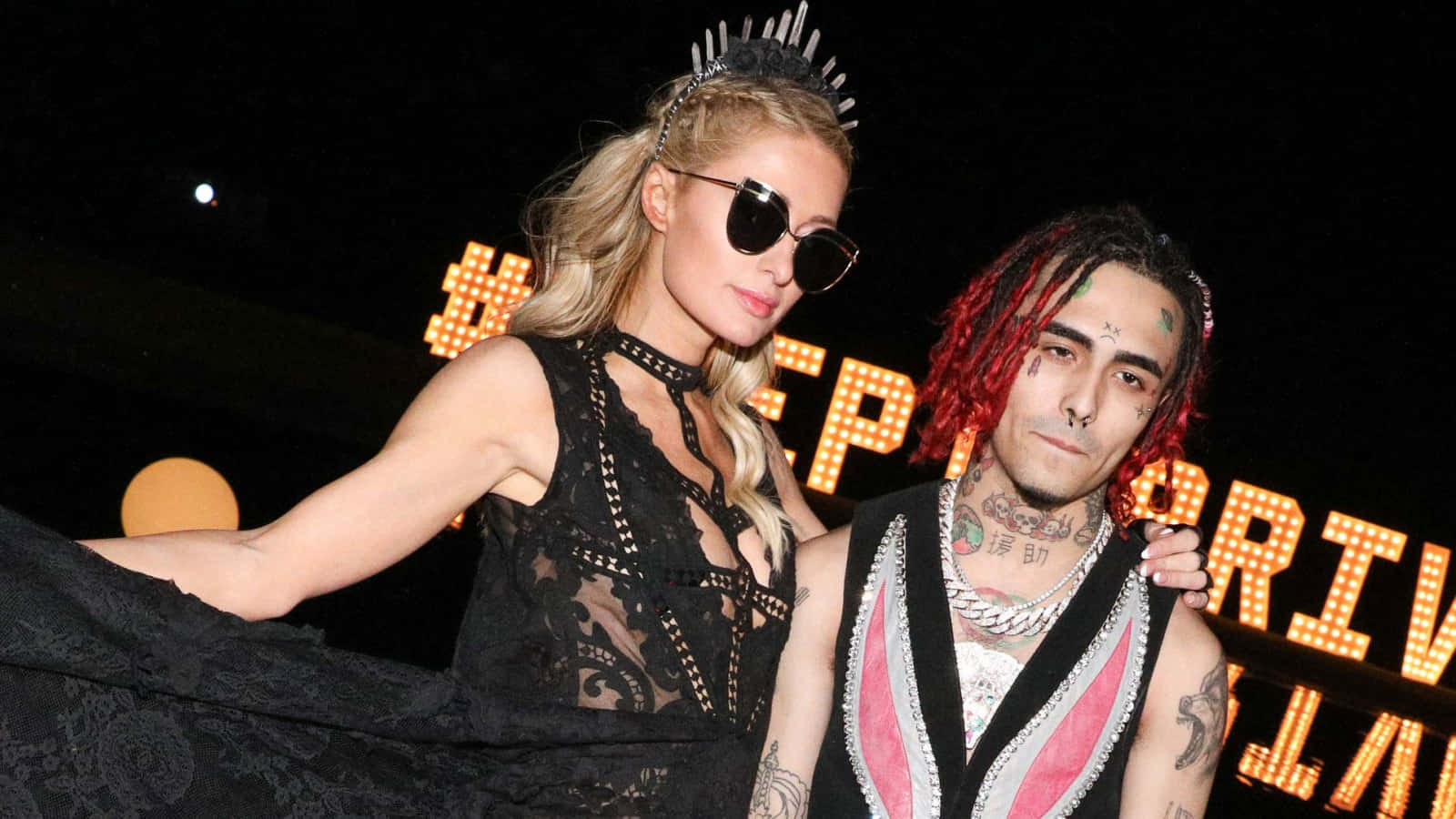桌布 壁紙 — Paris Hilton 與饒舌歌手 6ix9ine 在夜間活動中華麗擺姿勢，身後有發光的橙色標誌。