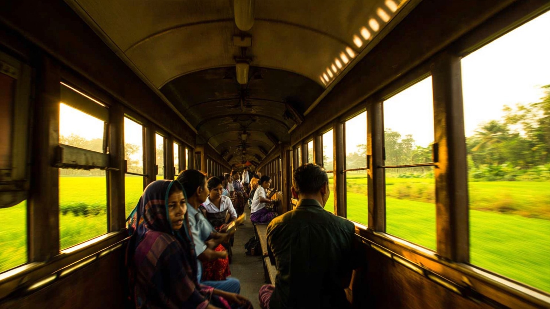 Yangon Circular Train Ride 壁紙 — 乘客坐在行駛中的火車車廂內，金色的陽光透過窗戶照射進來，顯露出外面模糊的綠色田野。
