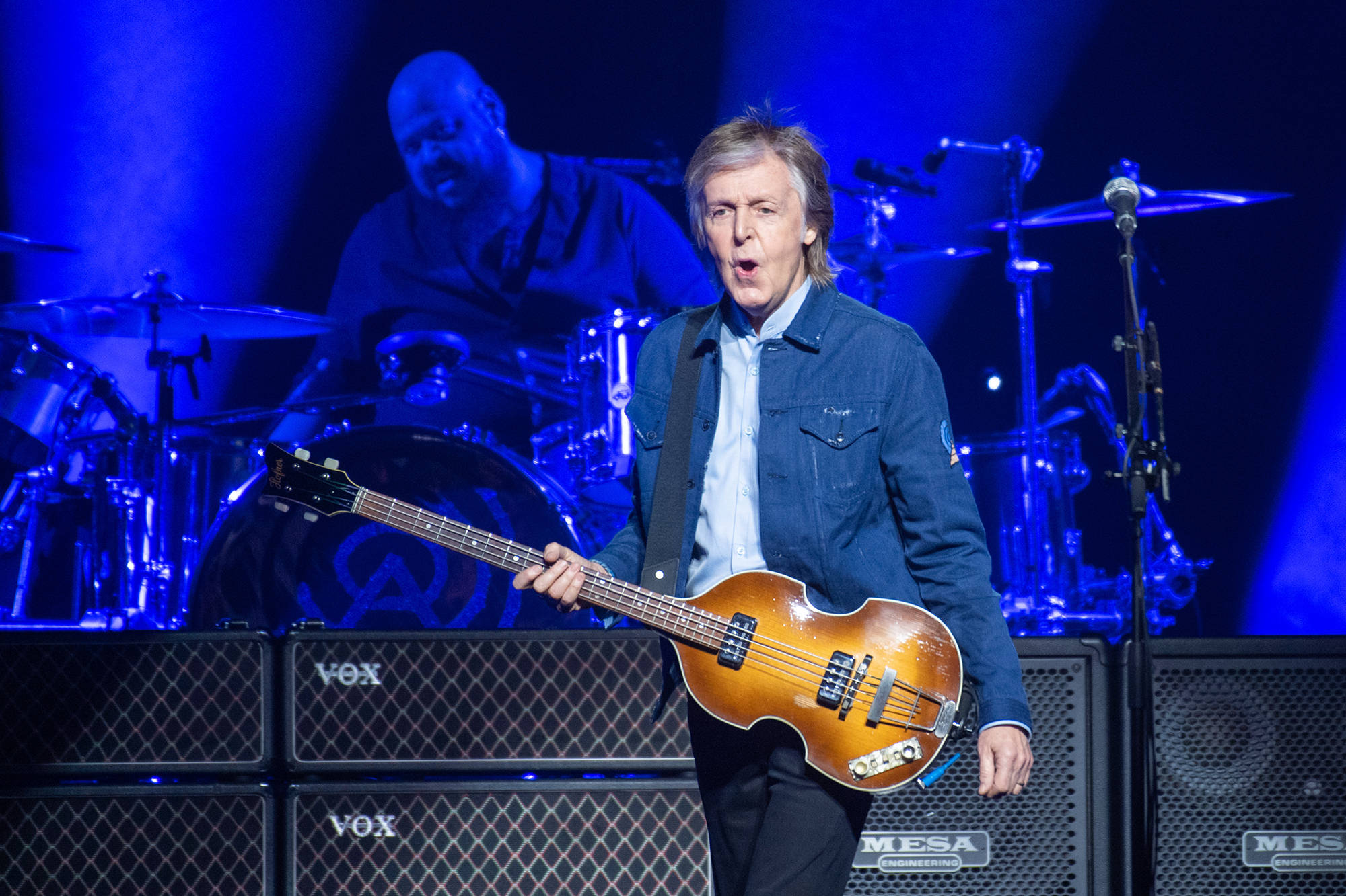 吉他動圖 桌布 壁紙 — Paul McCartney 在充滿活力的藍色舞台燈光下熱情地彈奏一把漸層色貝斯吉他，身後有一位鼓手。