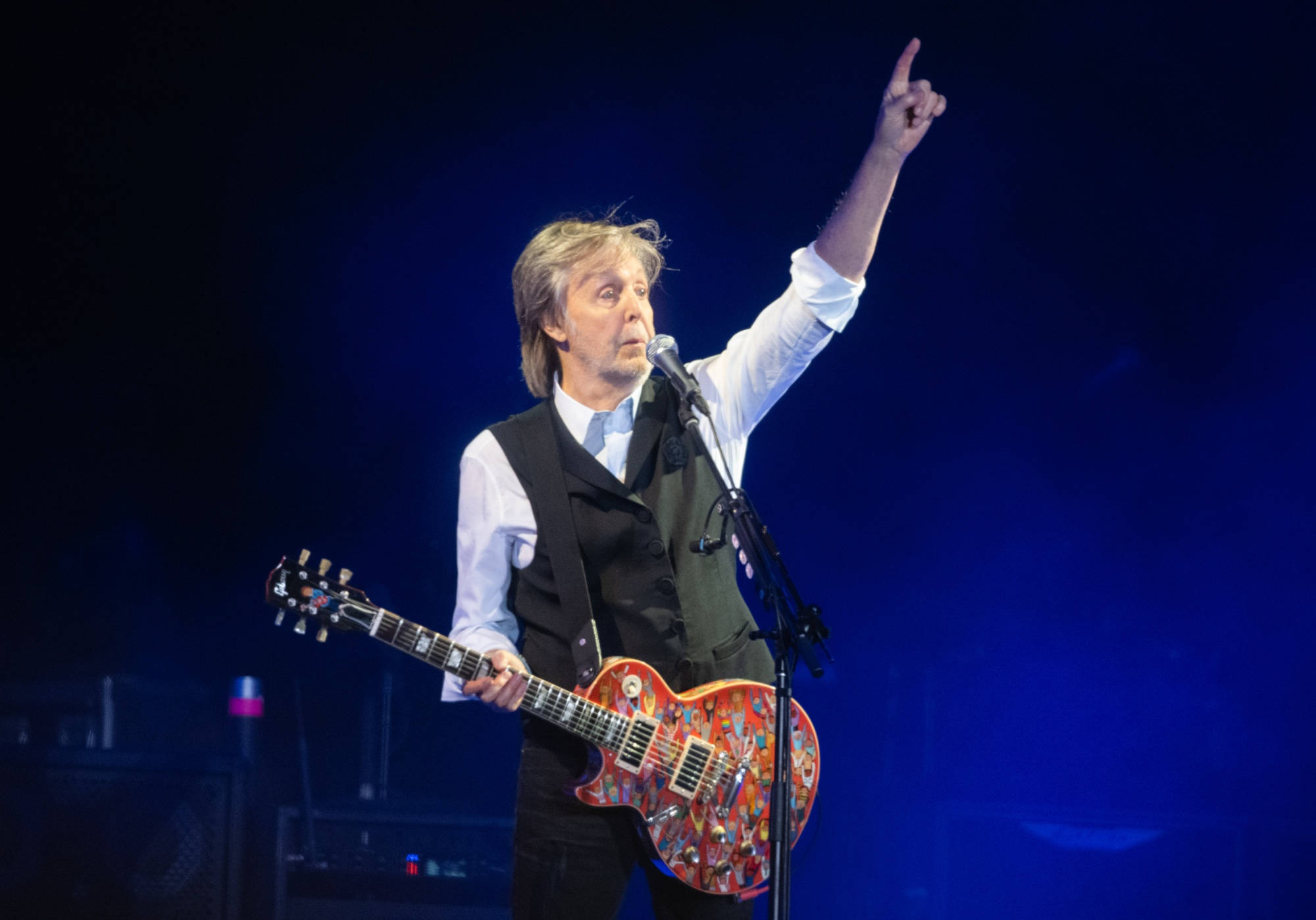 吉他動圖 桌布 壁紙 — Paul McCartney 在舞台上表演，手持色彩繽紛的吉他，在藍色舞台燈光下向上指。