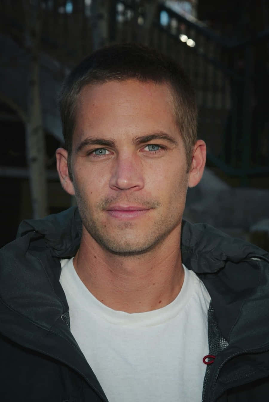 Paul Walker 桌布 壁紙 — Paul Walker 的特寫肖像，他有強烈的藍眼睛，穿著一件深色外套在白色T恤外面，背景是模糊的城市景觀。
