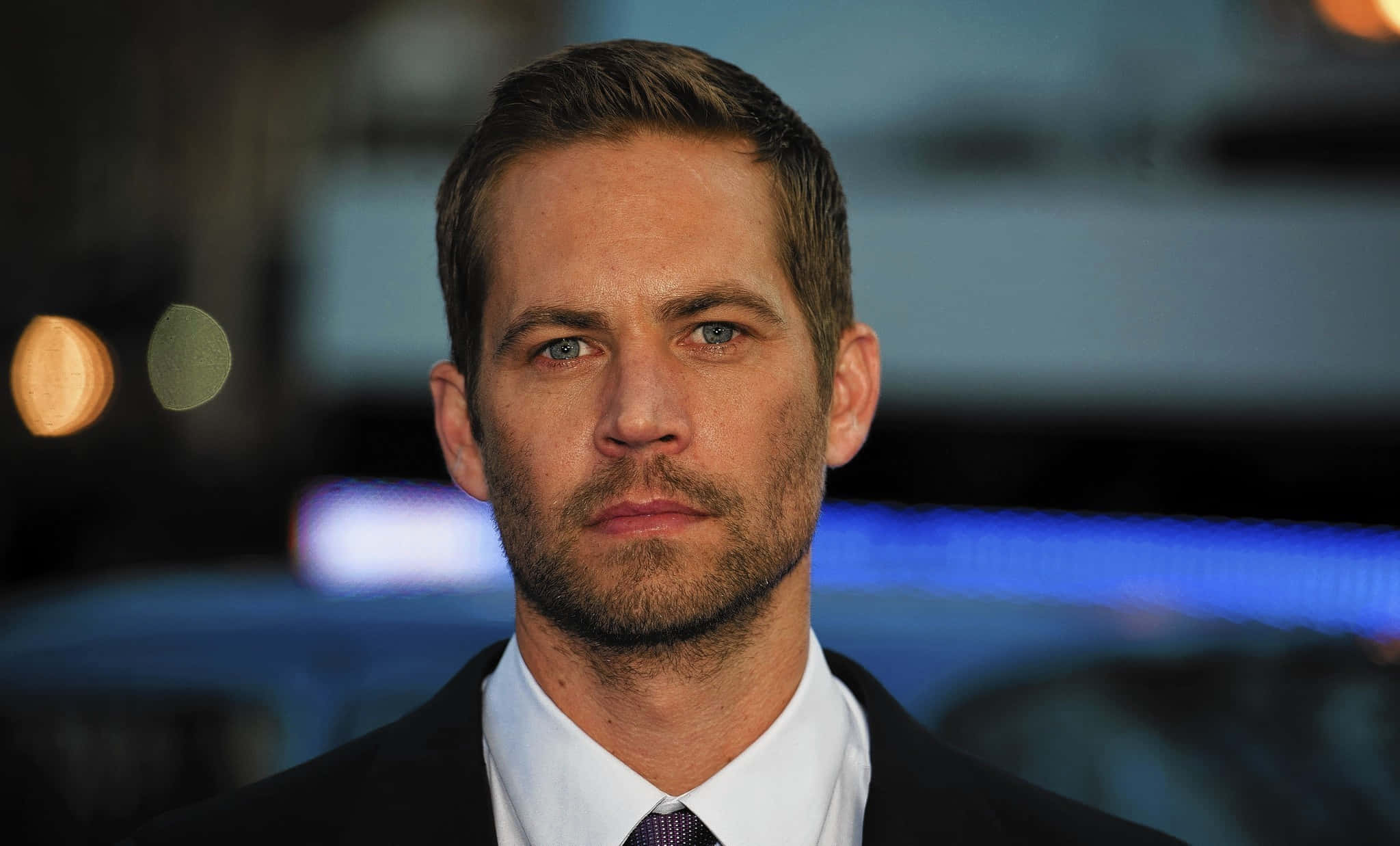 Paul Walker 桌布 壁紙 — Paul Walker 身穿深色西裝和領帶的特寫肖像，背景是模糊的城市夜景和散景燈光。