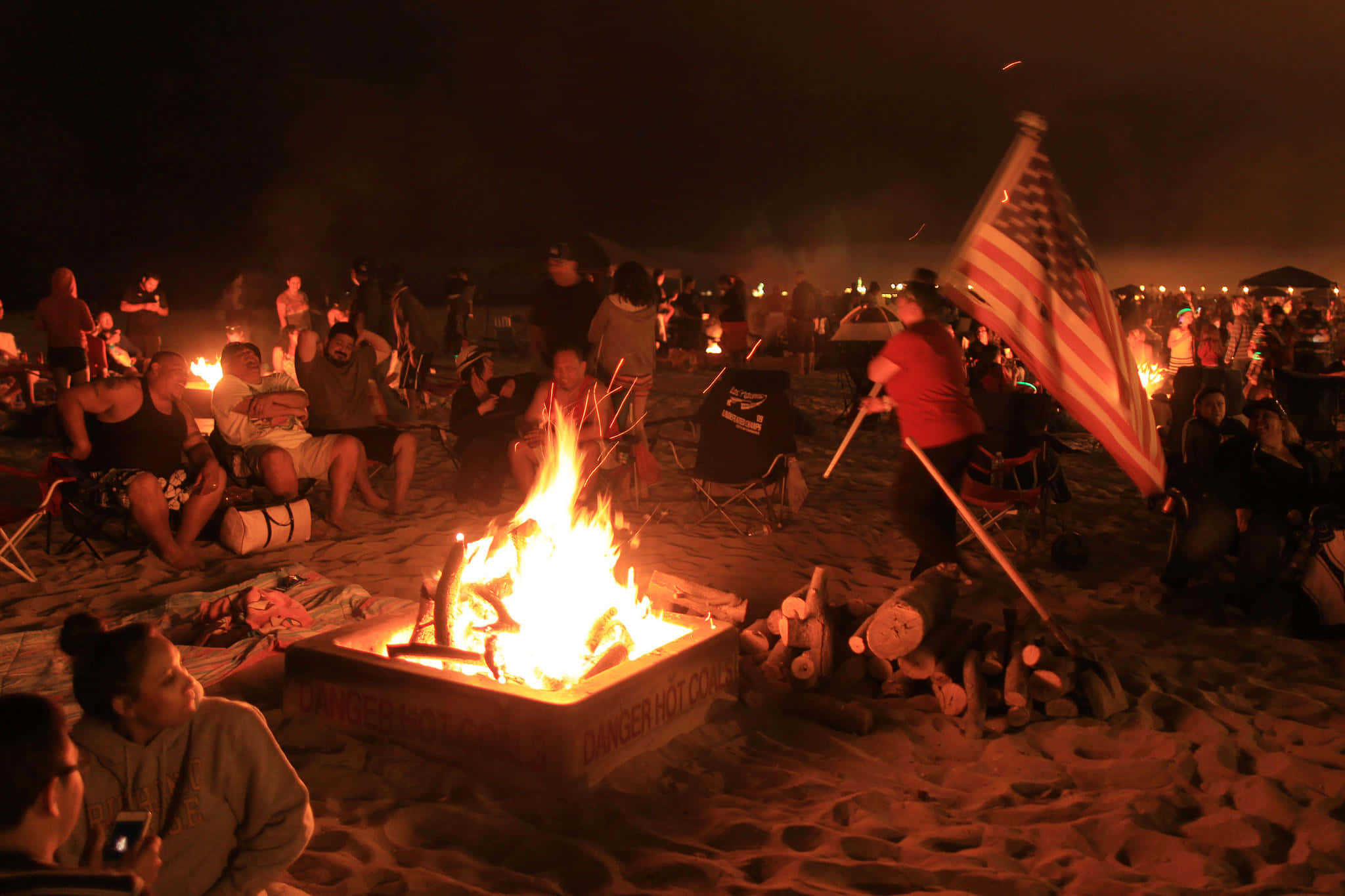 Beach Campfire 桌布 壁紙 — 人們在夜晚聚集在熊熊燃燒的海灘營火周圍，在黑暗的天空下揮舞美國國旗。