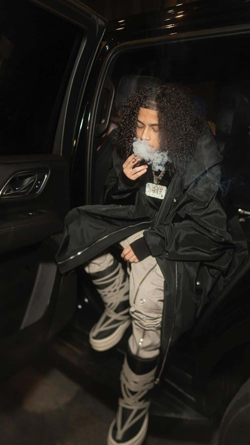  壁紙 — 一個有捲髮的人在夜晚坐在黑色車的打開的門上呼出煙霧。