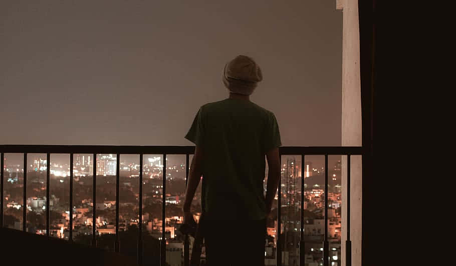 HD Aesthetic 桌布 壁紙 — 一個人戴著毛帽，夜晚獨自站在陽台上，俯瞰著被溫暖燈光照亮的廣闊城市景觀。