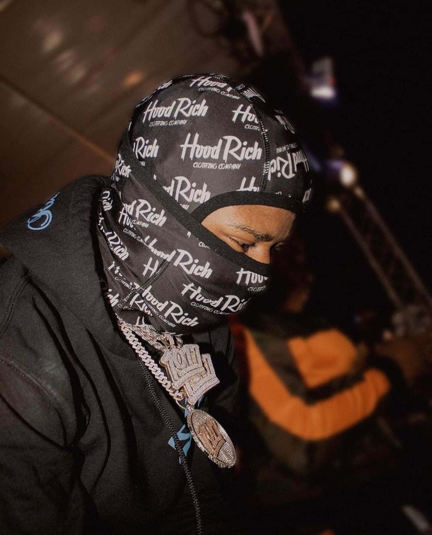  壁紙 — 一個戴著黑色 'Hood Rich' 頭巾和厚重銀鏈的人，在昏暗的城市環境中低頭看。