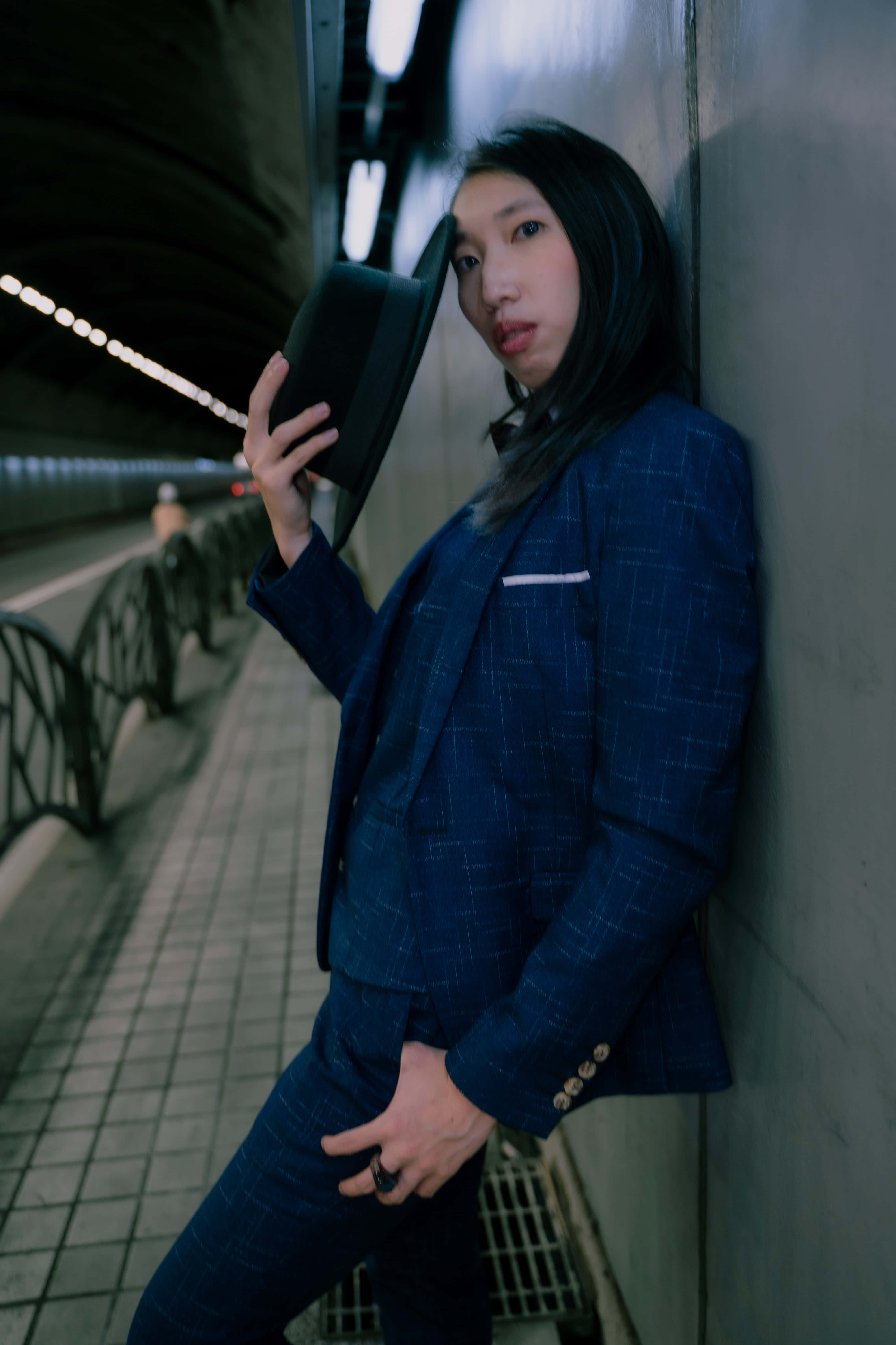 Navy Blue Blazer 桌布 壁紙 — 一個穿著藍色格紋西裝的人靠著隧道牆壁，在昏暗的人造燈光下拿著一頂高帽。