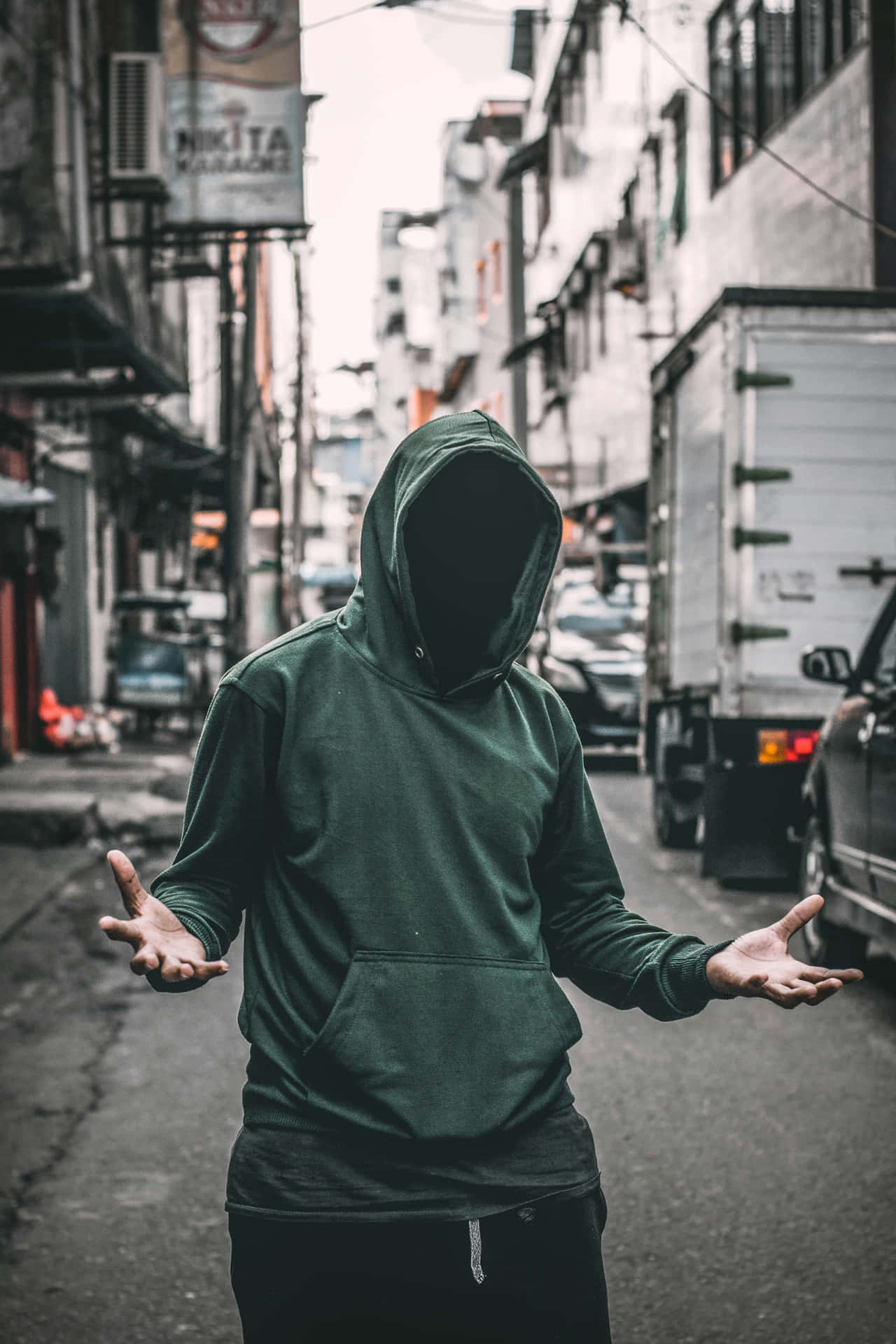  壁紙 — 一個身穿深綠色連帽衫、臉部隱藏的人，站在狹窄的城市小巷中，雙手向鏡頭張開。