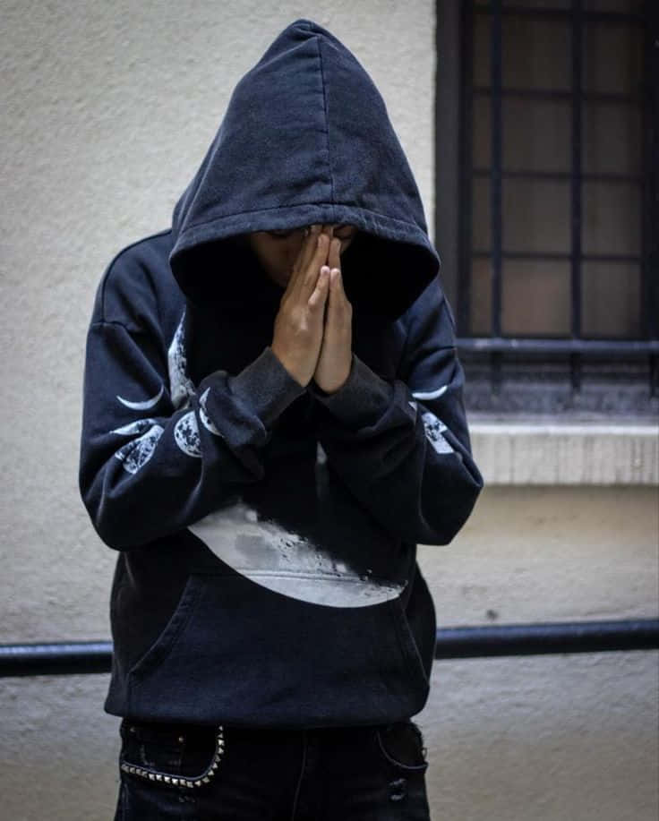  壁紙 — 一個穿著深色連帽衫的人，在帶有鐵欄杆的窗戶附近，用雙手遮住臉，靠著素牆。