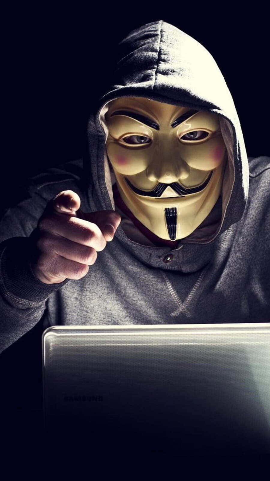  壁紙 — 一個穿著灰色連帽衫、戴著蓋伊·福克斯面具的人，在黑暗的房間裡指責地指著一台筆記型電腦。