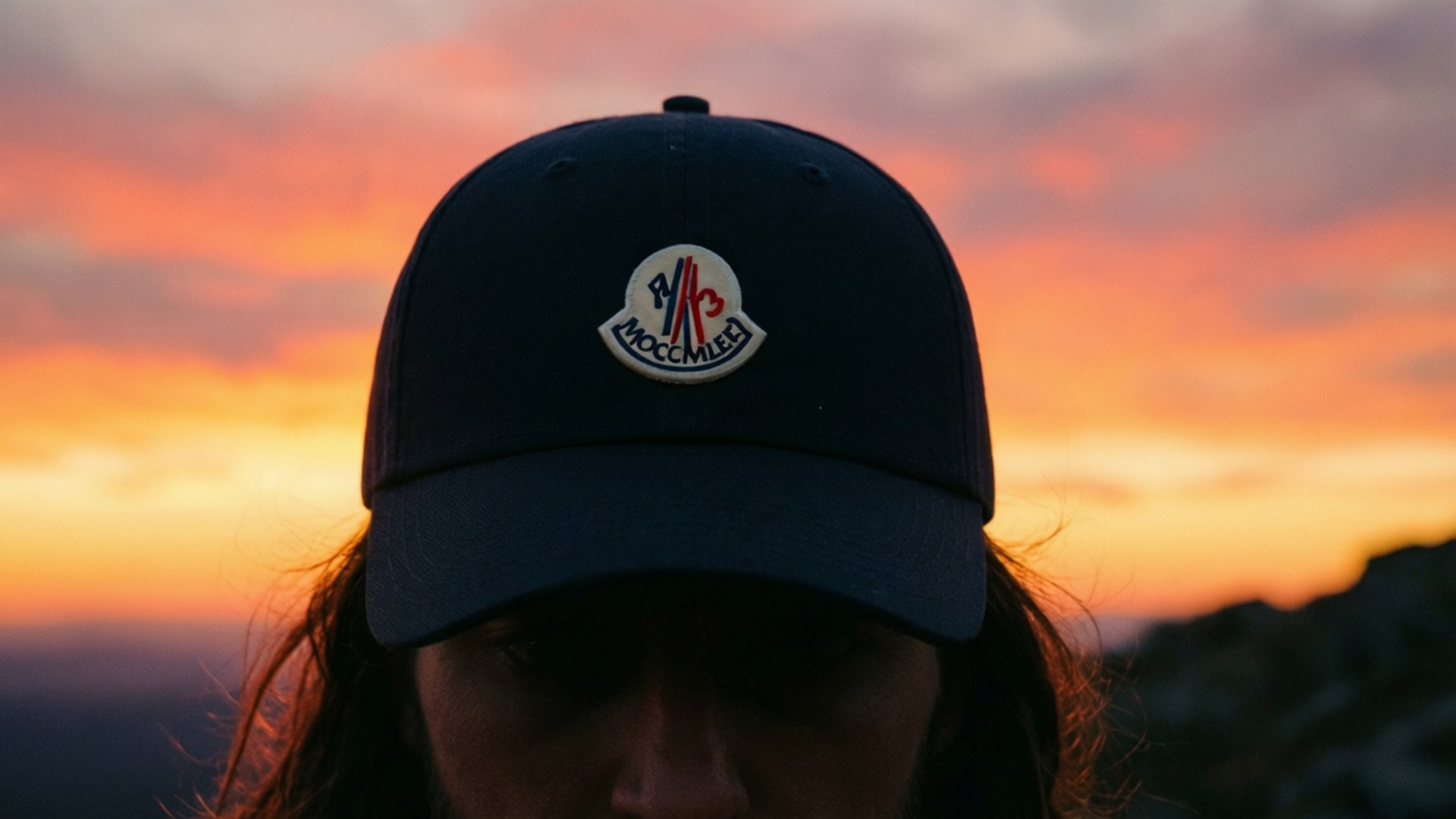 Moncler 桌布 壁紙 — 一個人戴著海軍藍 Moncler 帽，在熾熱的橙粉紅色日落天空下形成剪影。