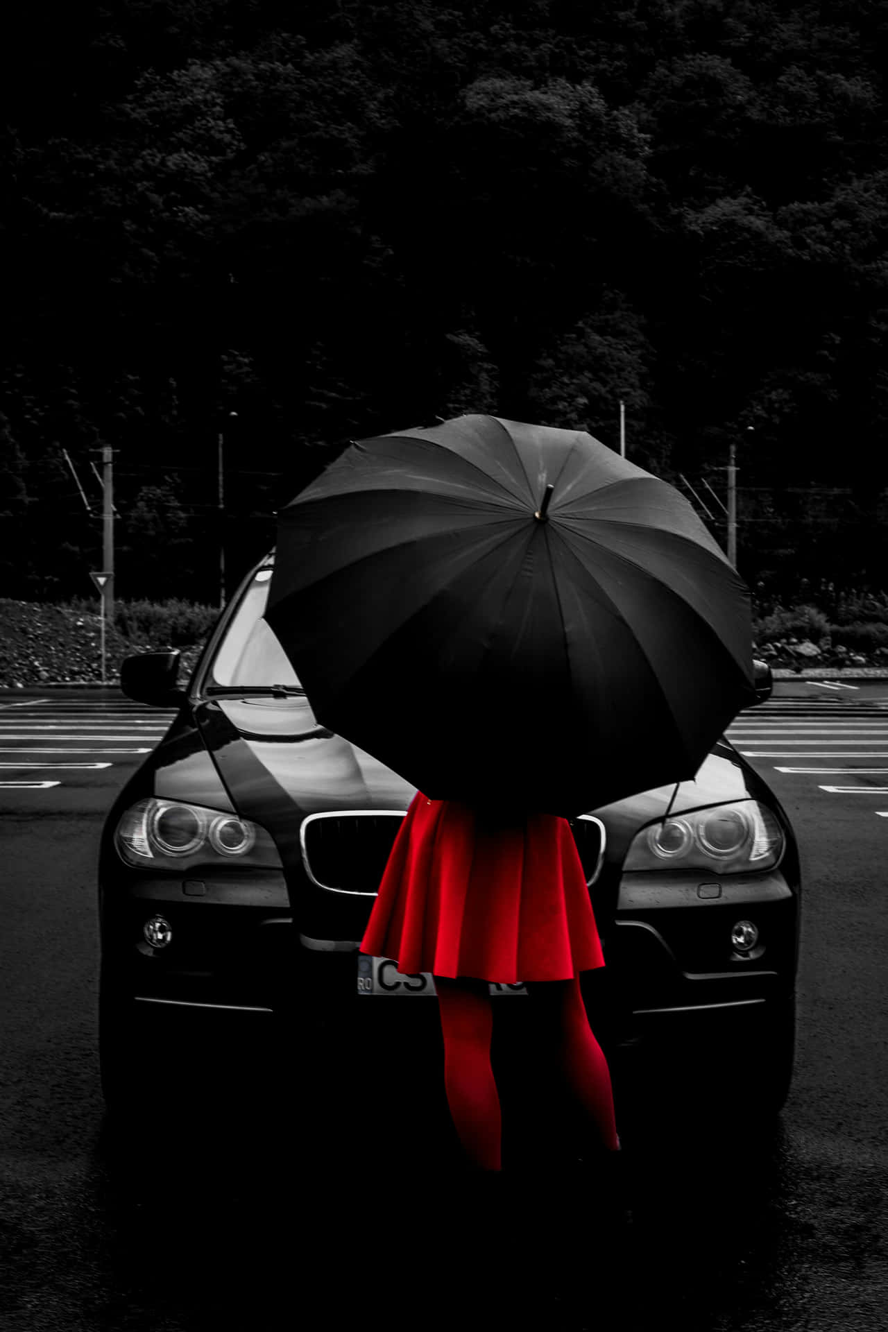  壁紙 — 一個穿著鮮紅色裙子的人，在雨中街道上，站在一輛黑色汽車前，手持一把黑色雨傘，背景有深色樹木。