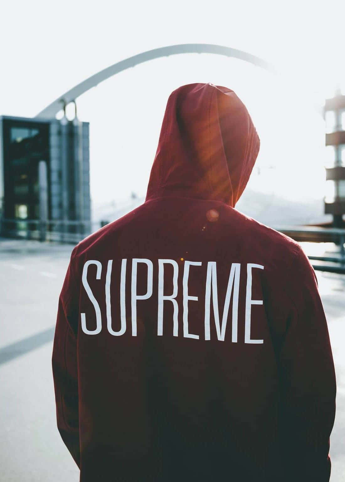  壁紙 — 一個穿著紅色連帽衫、背面印有「SUPREME」的人背對鏡頭，面向城市環境中的現代弧形拱門結構。