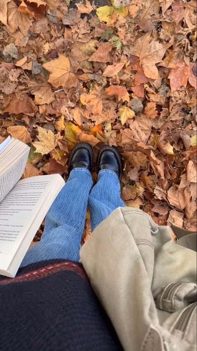  壁紙 — 一個人盤腿坐在金色秋葉地毯上閱讀一本書，穿著藍色牛仔褲和黑色鞋子。
