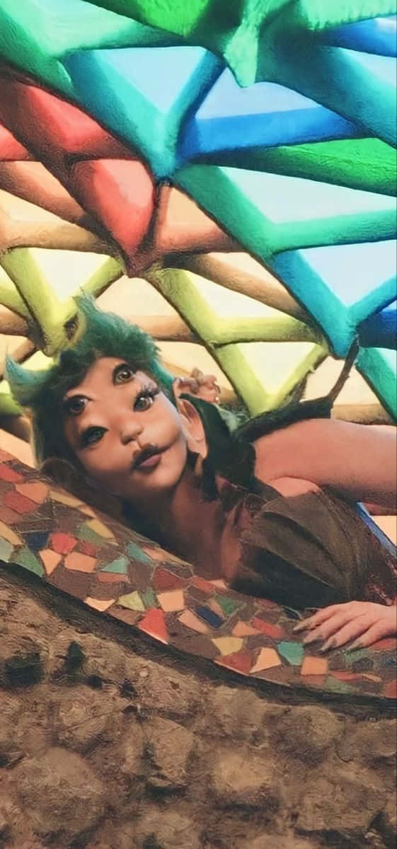 壁紙 — 一位擁有綠色頭髮和化妝的人躺在色彩繽紛的幾何馬賽克天篷下，身處質樸的泥土環境中。