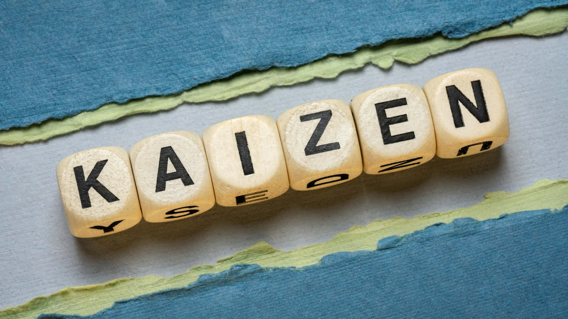  壁紙 — 拼出 kaizen 的木製字母方塊放在灰色表面上的撕破藍色和綠色紙上。