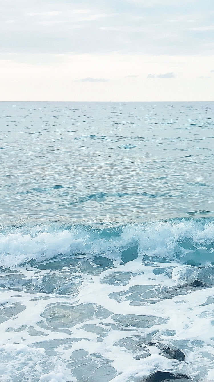  壁紙 — 平靜的海浪在柔和的多雲天空下，以柔和的藍白色調拍打著岩石海岸。
