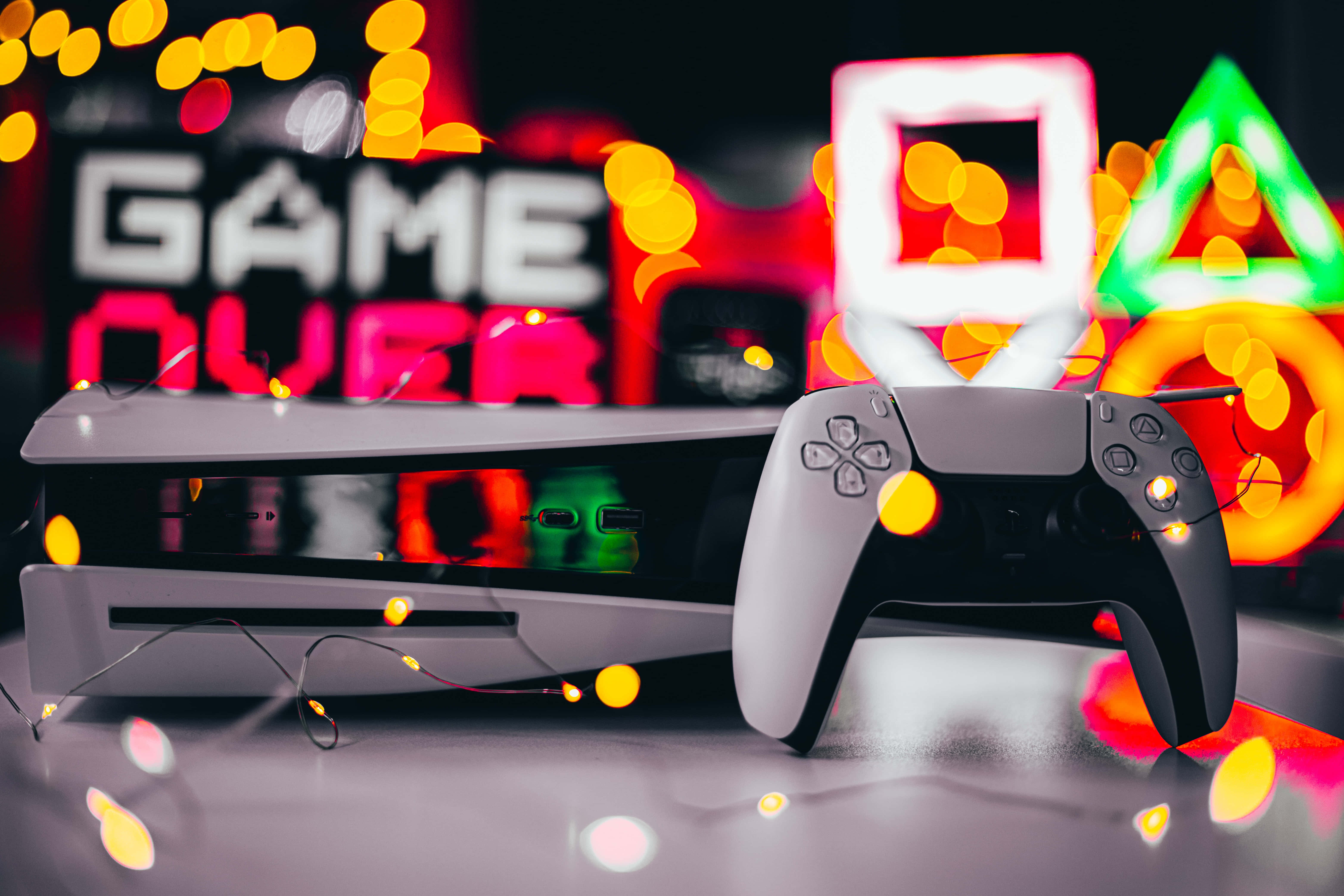 Gaming Consoles 桌布 壁紙 — PlayStation主機和控制器在彩色散景燈光下發光，背景有「GAME OVER」標誌。