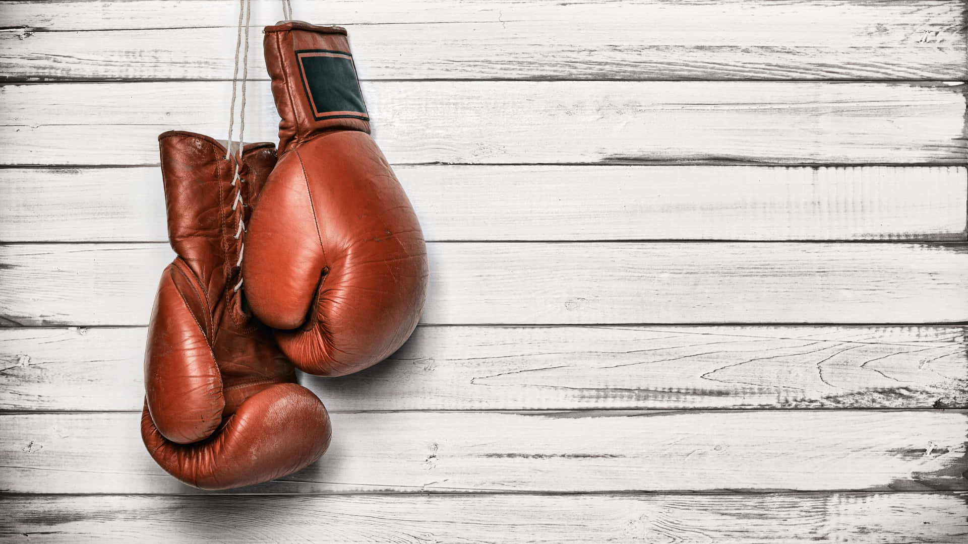  壁紙 — 破舊的棕色皮革拳擊手套用繩子掛在風化的白色木板背景上。