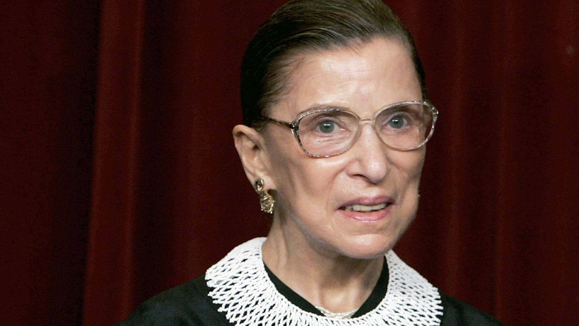 露絲·拜德·金斯伯格 桌布 壁紙 — Ruth Bader Ginsburg 身穿法官袍，以深紅色窗簾為背景的肖像。