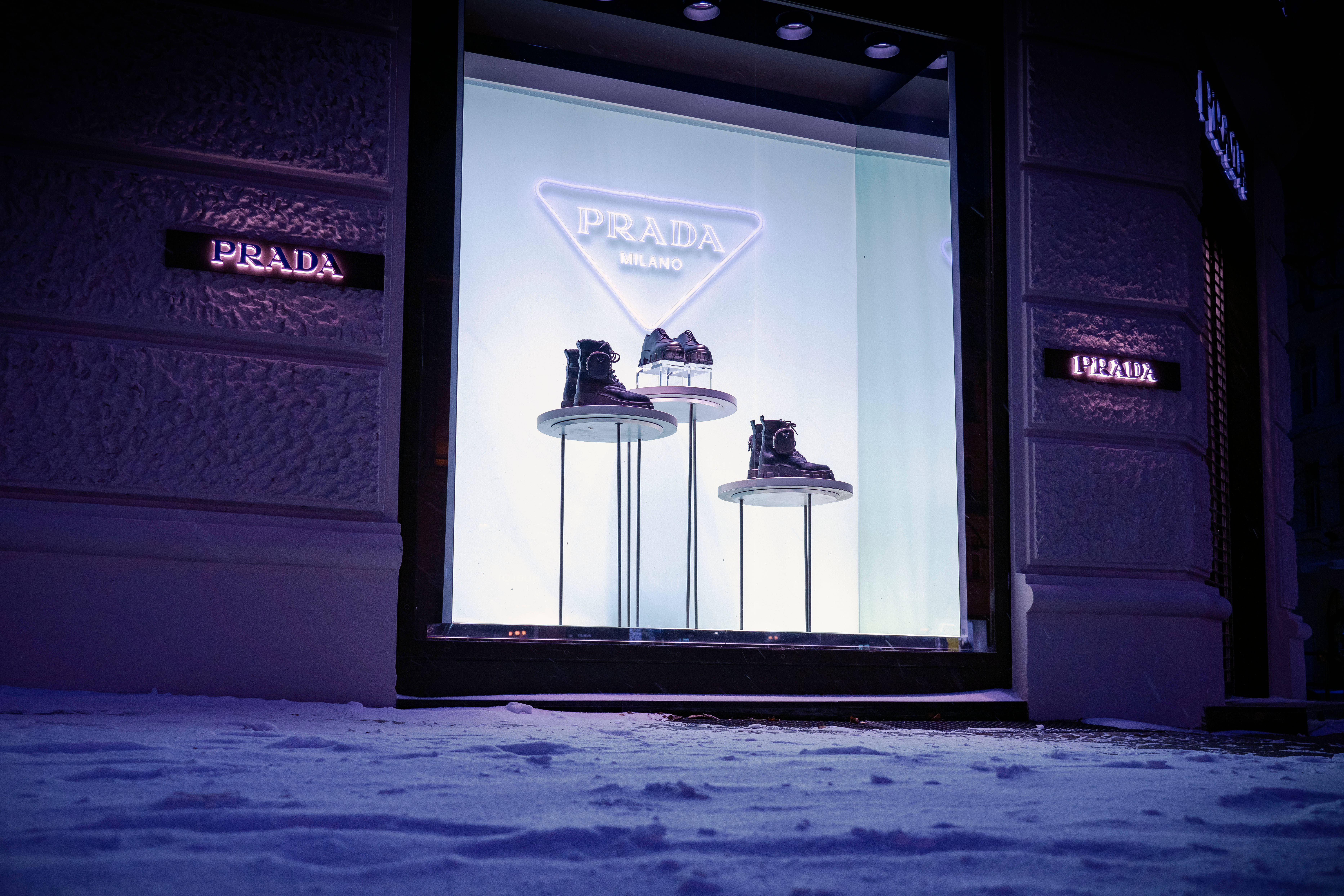 Prada 桌布 壁紙 — Prada 商店櫥窗在雪夜城市背景下，以霓虹燈照亮展示台上的鞋子。