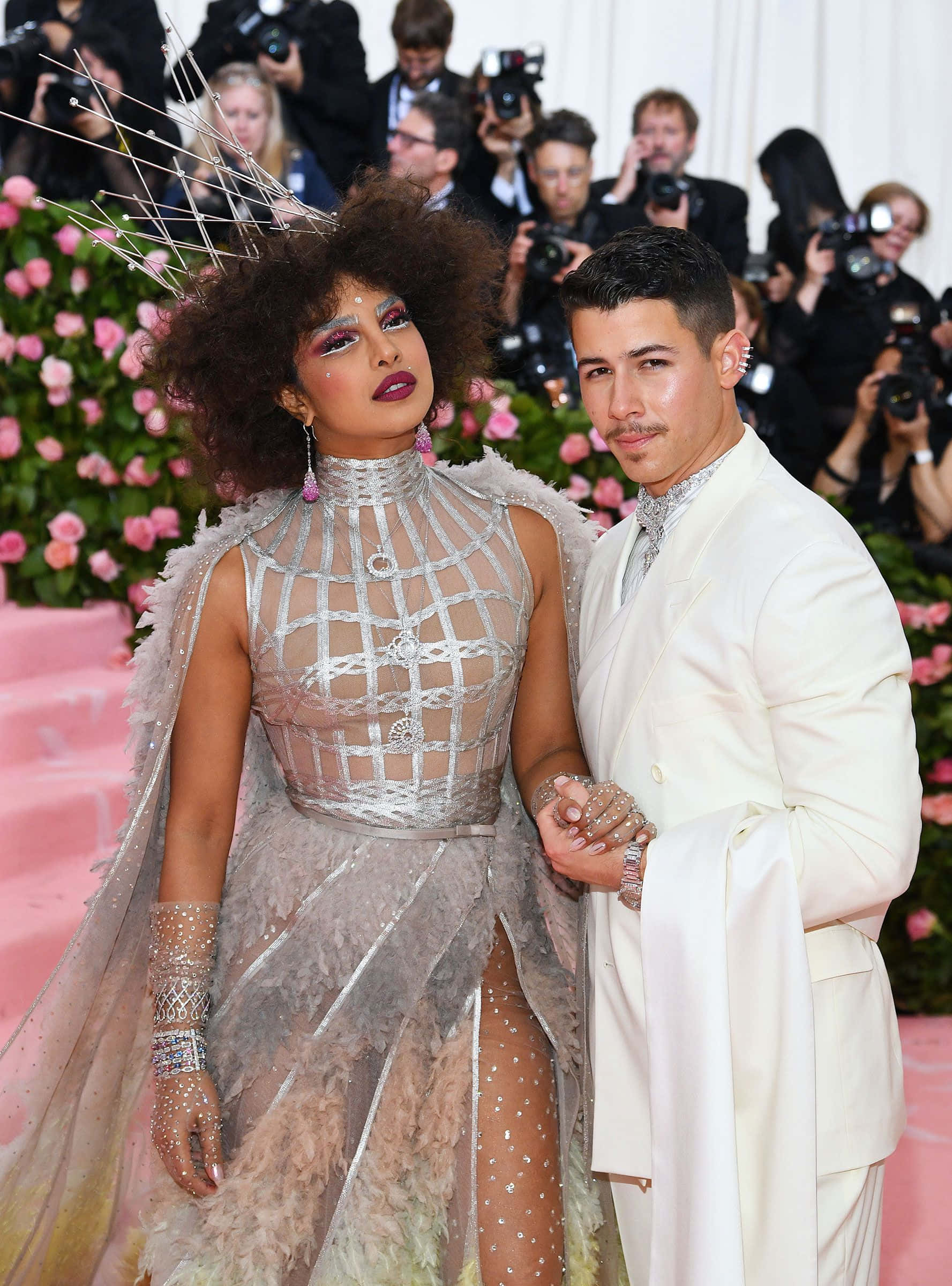 Met Gala 桌布 壁紙 — Priyanka Chopra 身穿銀色網格禮服，搭配羽毛披肩和金屬皇冠，站在身穿白色西裝的 Nick Jonas 旁邊，在粉紅色地毯上，周圍有攝影師和粉紅色玫瑰。