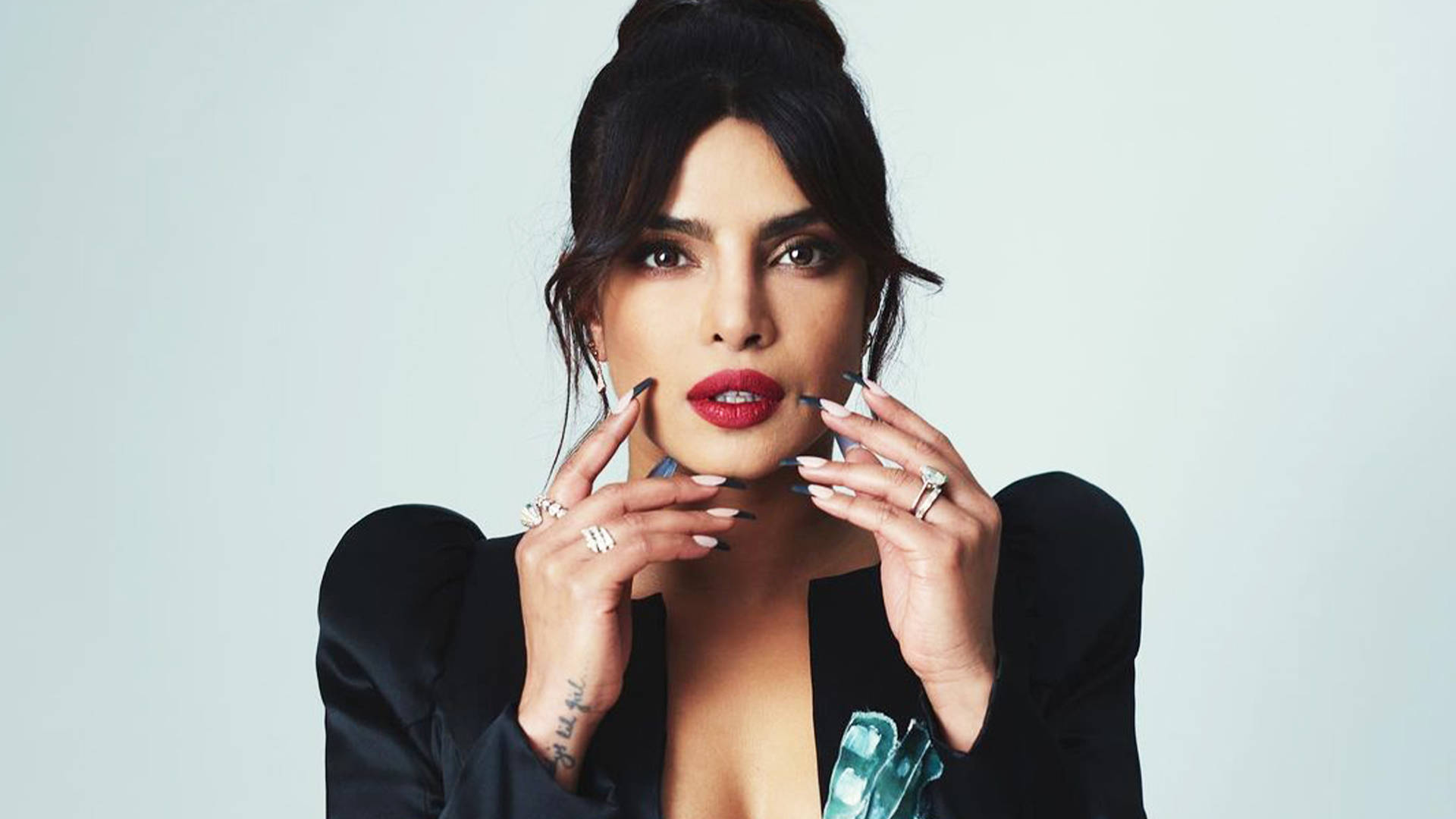 Priyanka 桌布 壁紙 — Priyanka Chopra 自信地擺姿勢，雙手靠近臉部，背景為純淺灰色，身穿黑色西裝外套和紅色唇膏。