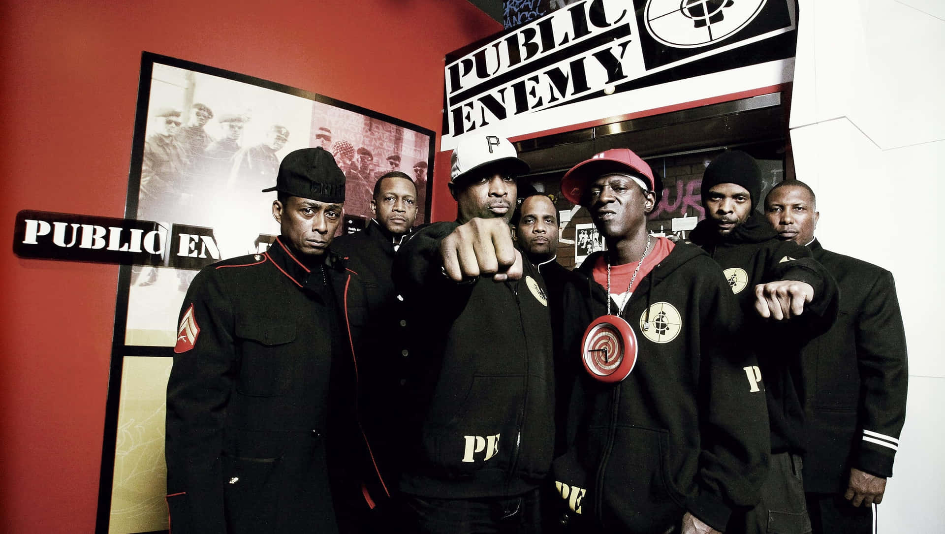  壁紙 — Public Enemy 樂團成員身穿帶有紅色點綴的黑色外套，在他們標誌性的標誌和裱框照片前挑釁地擺姿勢。