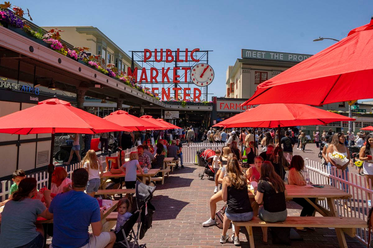 Pike Place Market 桌布 壁紙 — Public Market Center 充滿活力的戶外座位區，紅色遮陽傘下，人群熙攘，天空湛藍。
