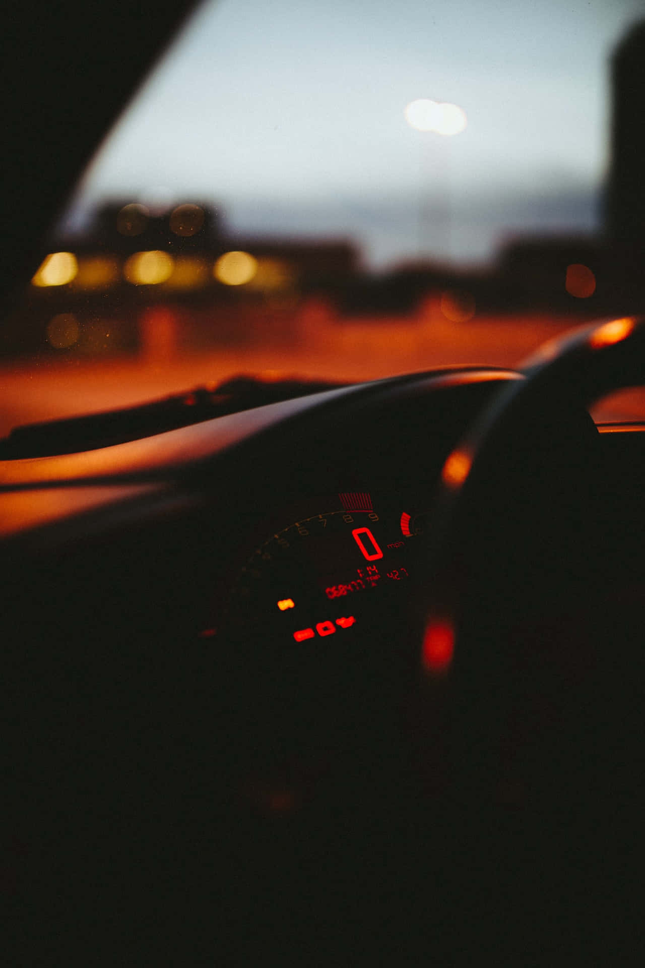 汽車儀表板在黃昏時亮起紅光，背後有模糊的城市燈光。 — 車內 壁紙