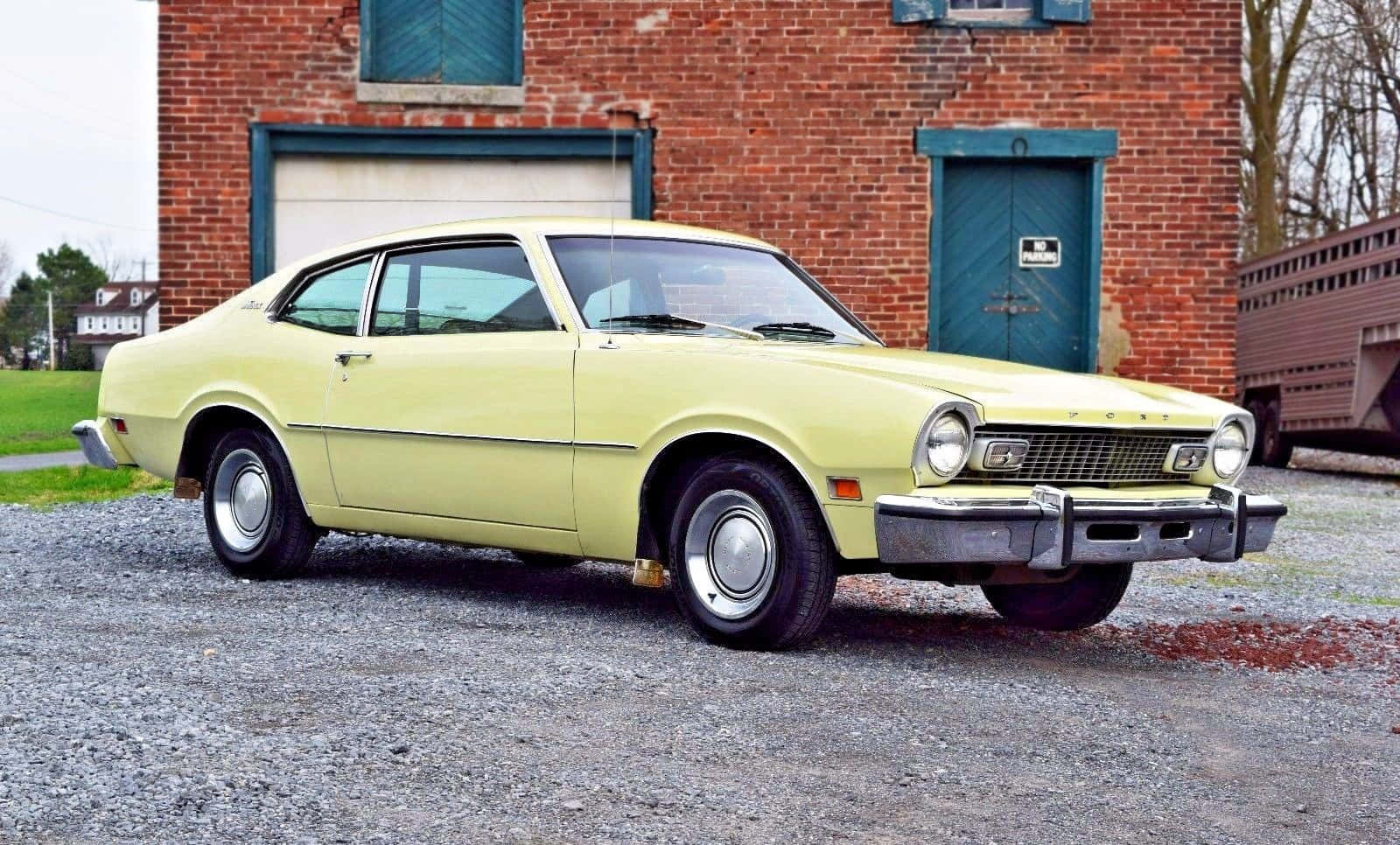 Ford Maverick 桌布 壁紙 — 一輛淺黃色經典福特 Maverick 雙門轎車停在碎石路上，前方是帶有藍色門的紅磚建築。