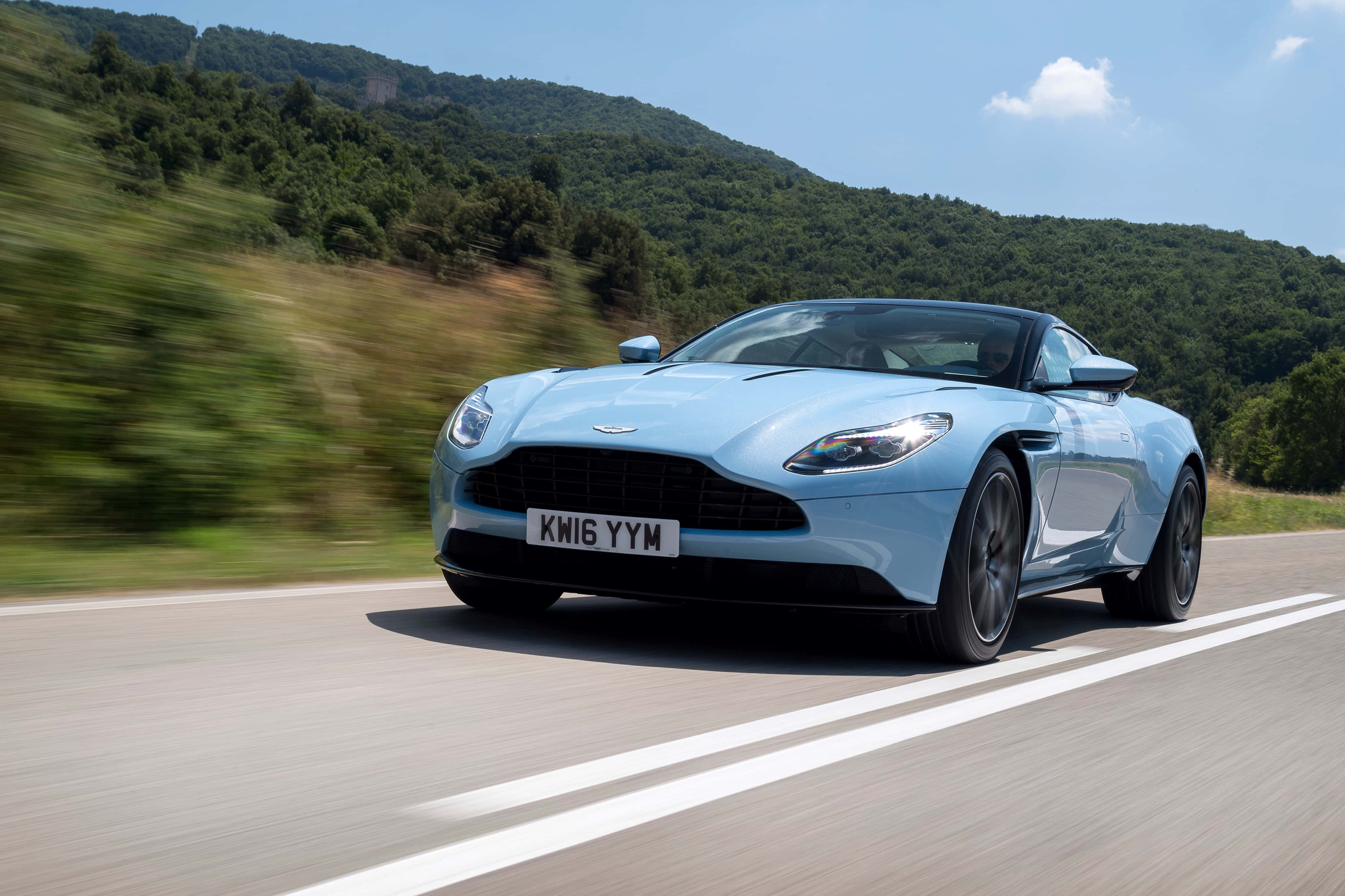 Aston Martin Db11 桌布 壁紙 — 一輛淺藍色Aston Martin跑車在晴朗藍天下，沿著蜿蜒的道路穿過翠綠的山丘疾馳。