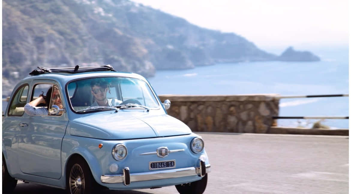 Fiat 500 桌布 壁紙 — 一輛淺藍色復古飛雅特500沿著海岸懸崖道路行駛，一名男子駕駛，一名女子從乘客座位揮手。