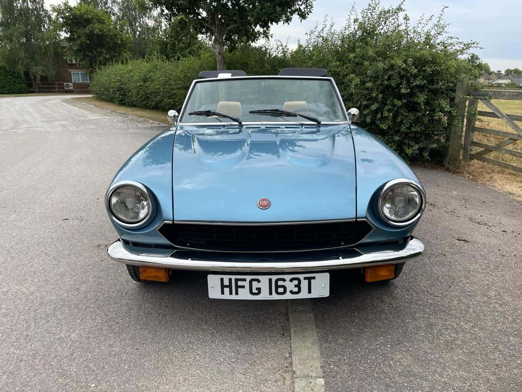 Fiat 124 Spider 桌布 壁紙 — 一架淺藍色經典菲亞特124 Spider敞篷車停泊在碎石車道上，背景有綠色籬笆和木門。