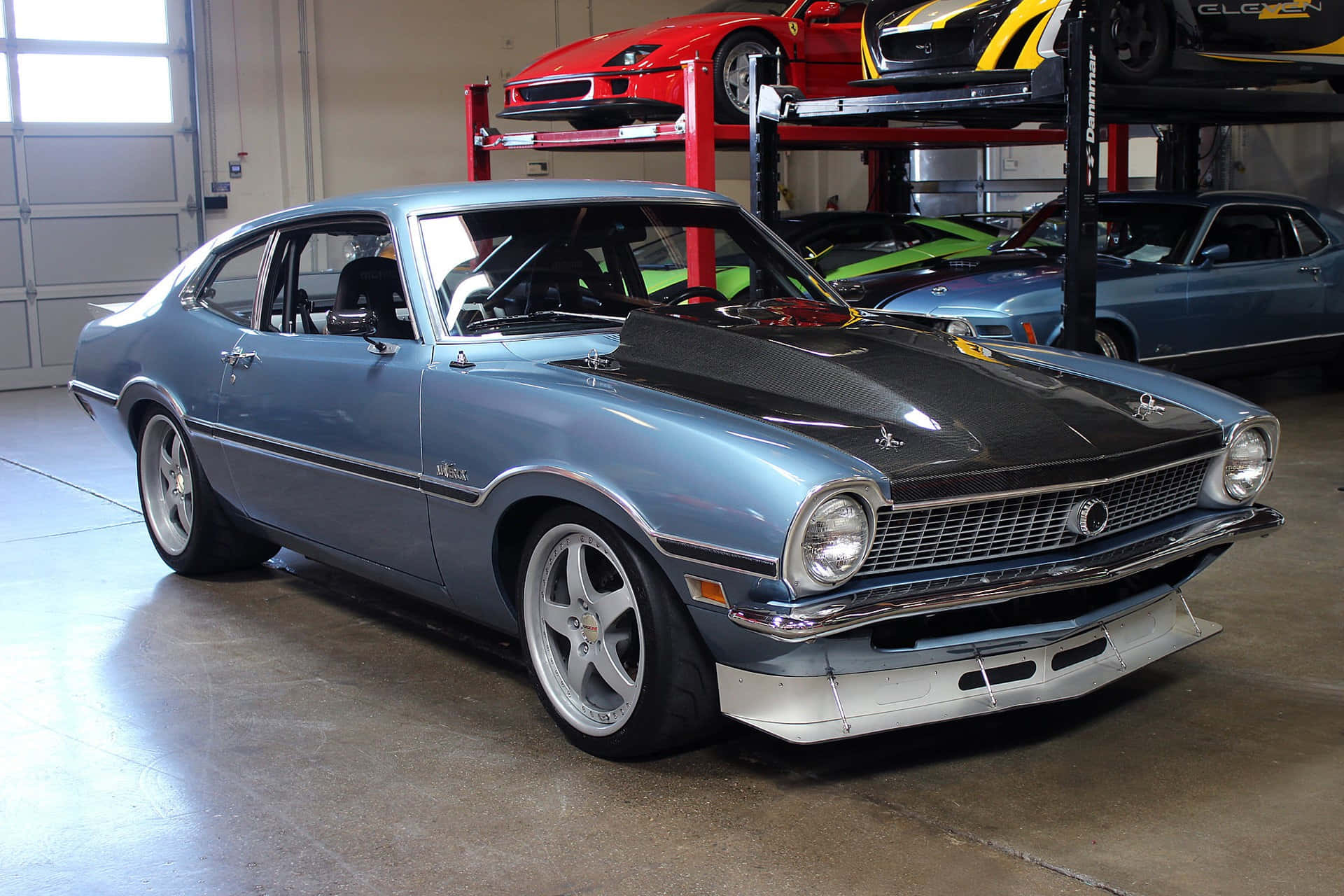 Ford Maverick 桌布 壁紙 — 一輛淺藍色經典肌肉車搭配黑色引擎蓋，在光線充足的車庫中顯眼地停放，周圍有其他復古和現代跑車在升降機上。