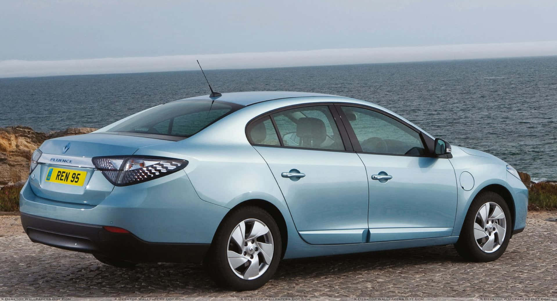 Renault Fluence 桌布 壁紙 — 一架淺藍色雷諾Fluence房車停泊在鵝卵石懸崖上，俯瞰著多雲天空下平靜的海洋。