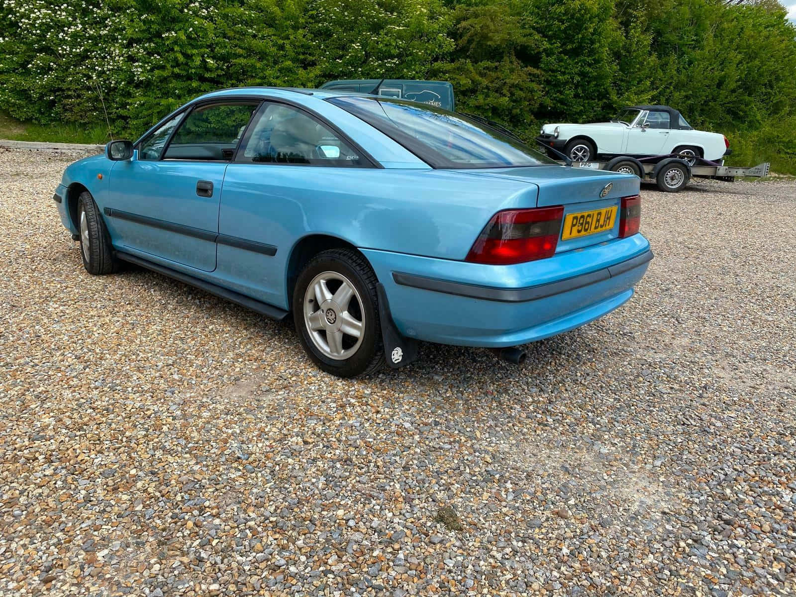 Opel Calibra 桌布 壁紙 — 一輛淺藍色雙門跑車停泊在碎石地上，後面有綠色樹木，附近拖車上有一輛白色經典汽車。