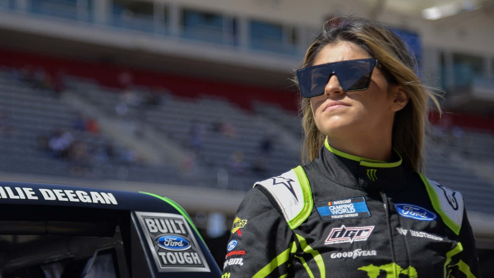  壁紙 — 賽車手Danica Patrick自信地站在她的福特NASCAR賽車旁邊，身後是陽光普照的體育場和空無一人的看台。