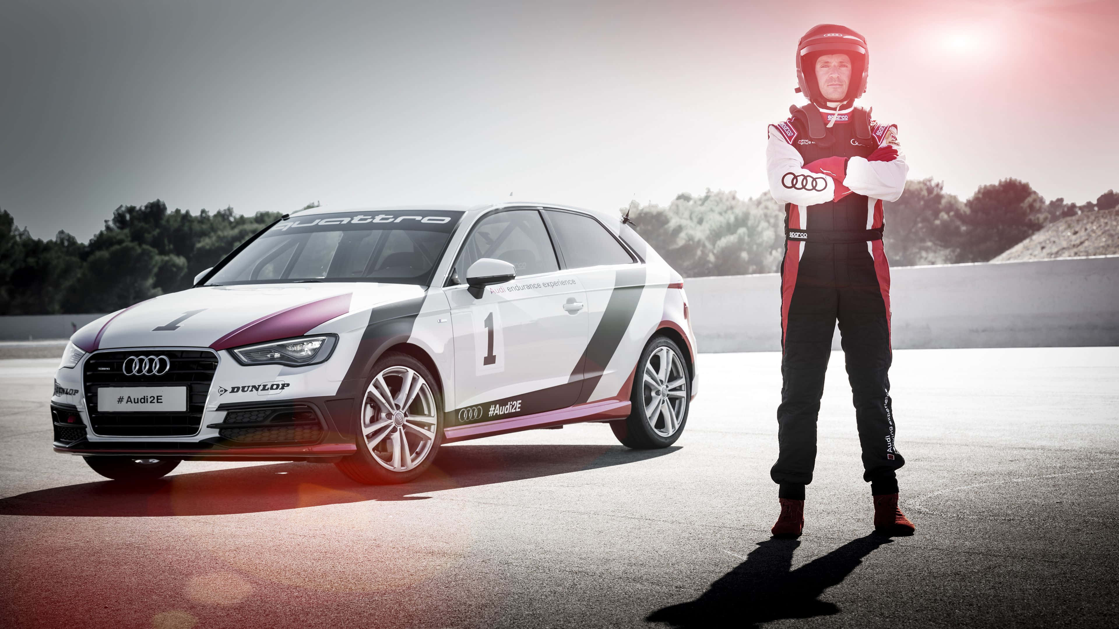 Audi A3 桌布 壁紙 — 一名身穿全套裝備的賽車手自信地站在白色奧迪賽車旁，背景是陽光明媚的賽道和樹木。