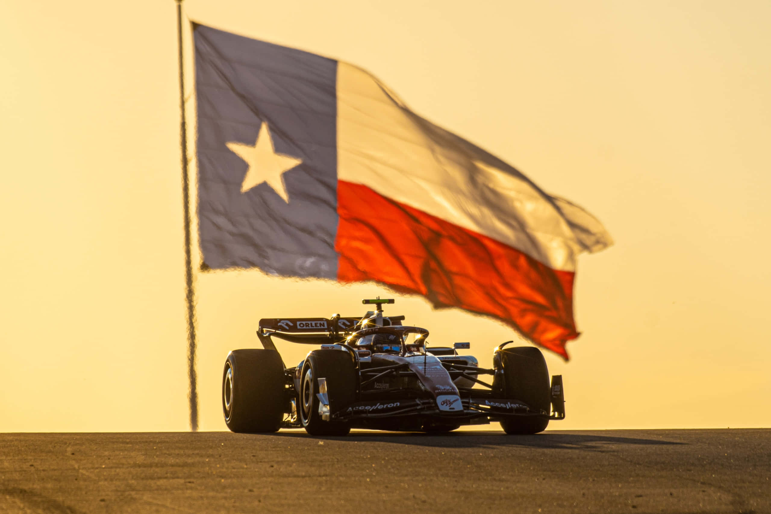 Formula 1 桌布 壁紙 — 一架時尚黑色賽車在金色夕陽天空下，疾馳掠過飄揚的德克薩斯州旗。