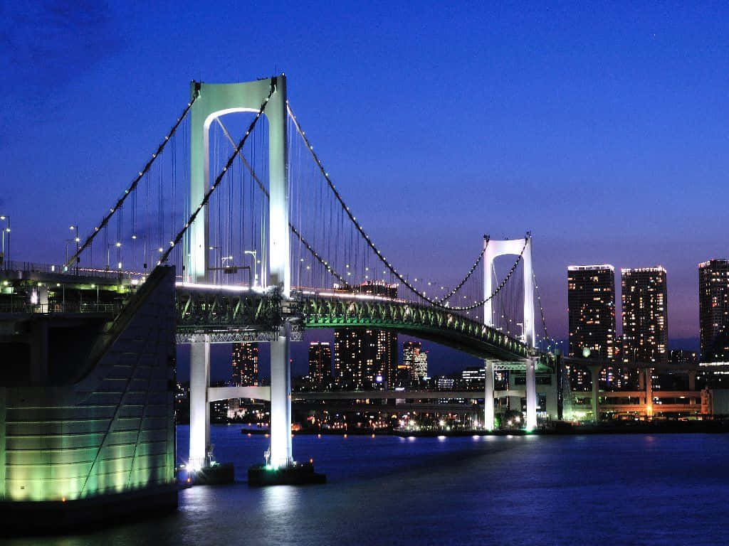 Rainbow Bridge 桌布 壁紙 — Rainbow Bridge 在黃昏時亮起，發出綠色光芒，倒映在水面上，以東京天際線為背景。