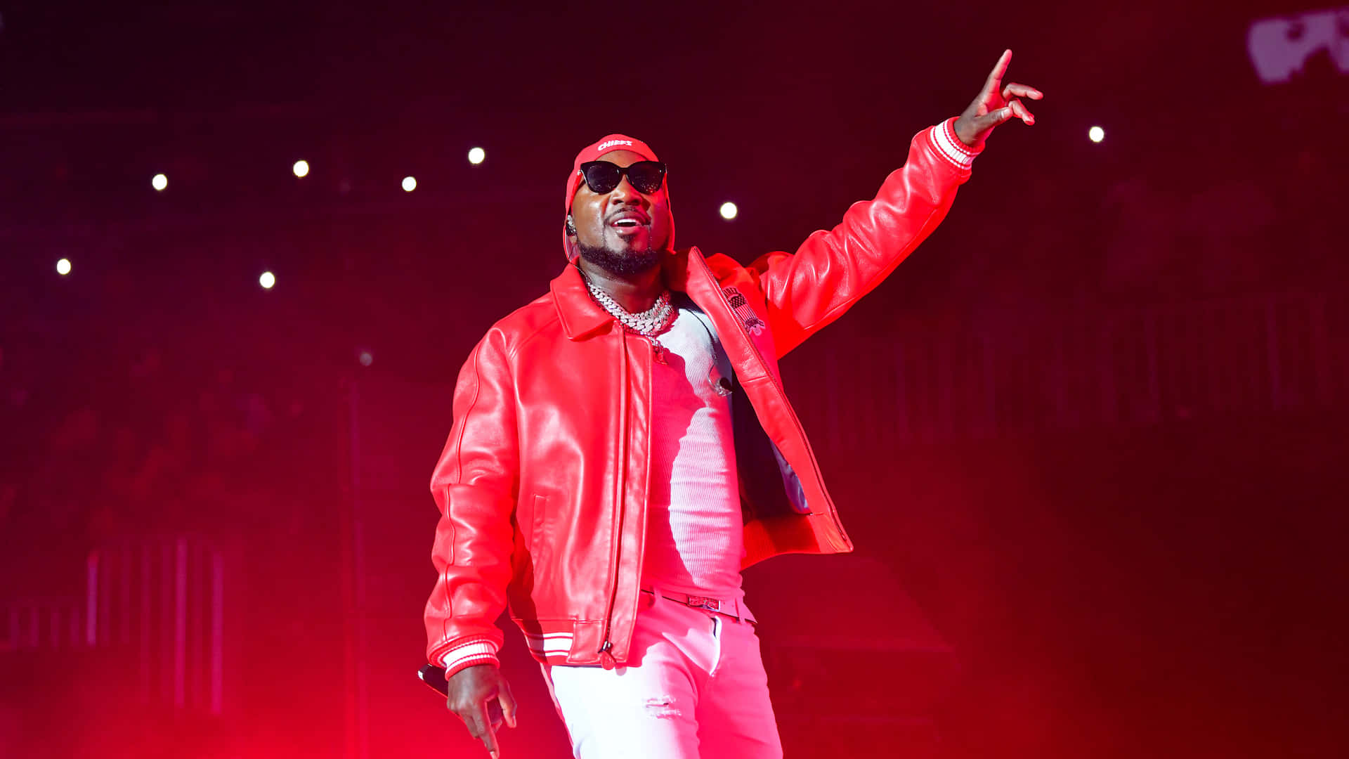  壁紙 — 一位身穿紅色皮夾克和白色褲子的饒舌歌手，在紅光舞台高舉手臂、戴著太陽眼鏡，充滿活力地表演。
