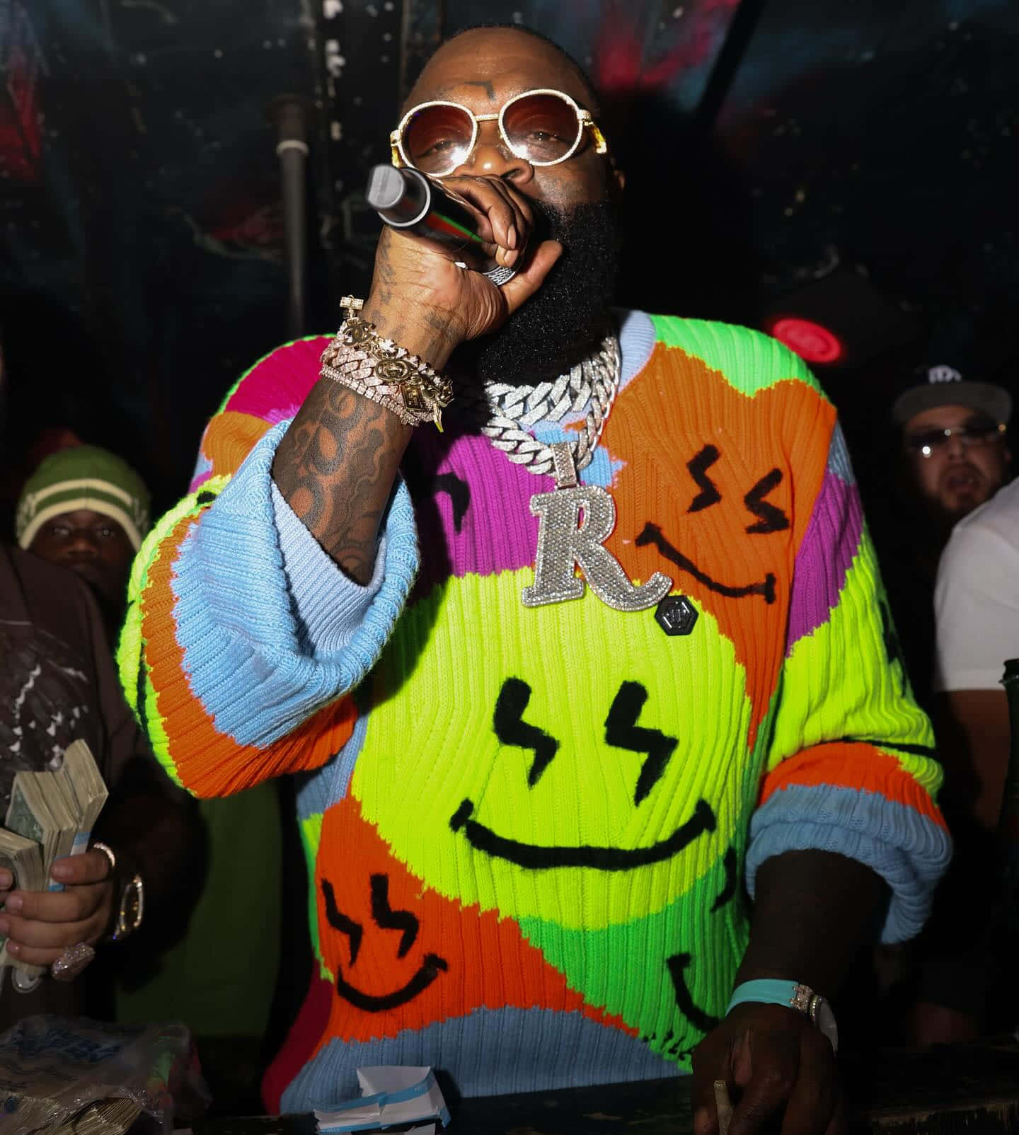  壁紙 — 饒舌歌手 Rick Ross 穿著一件色彩繽紛、帶有笑臉和閃電圖案的毛衣，在昏暗的活動中手持麥克風表演。
