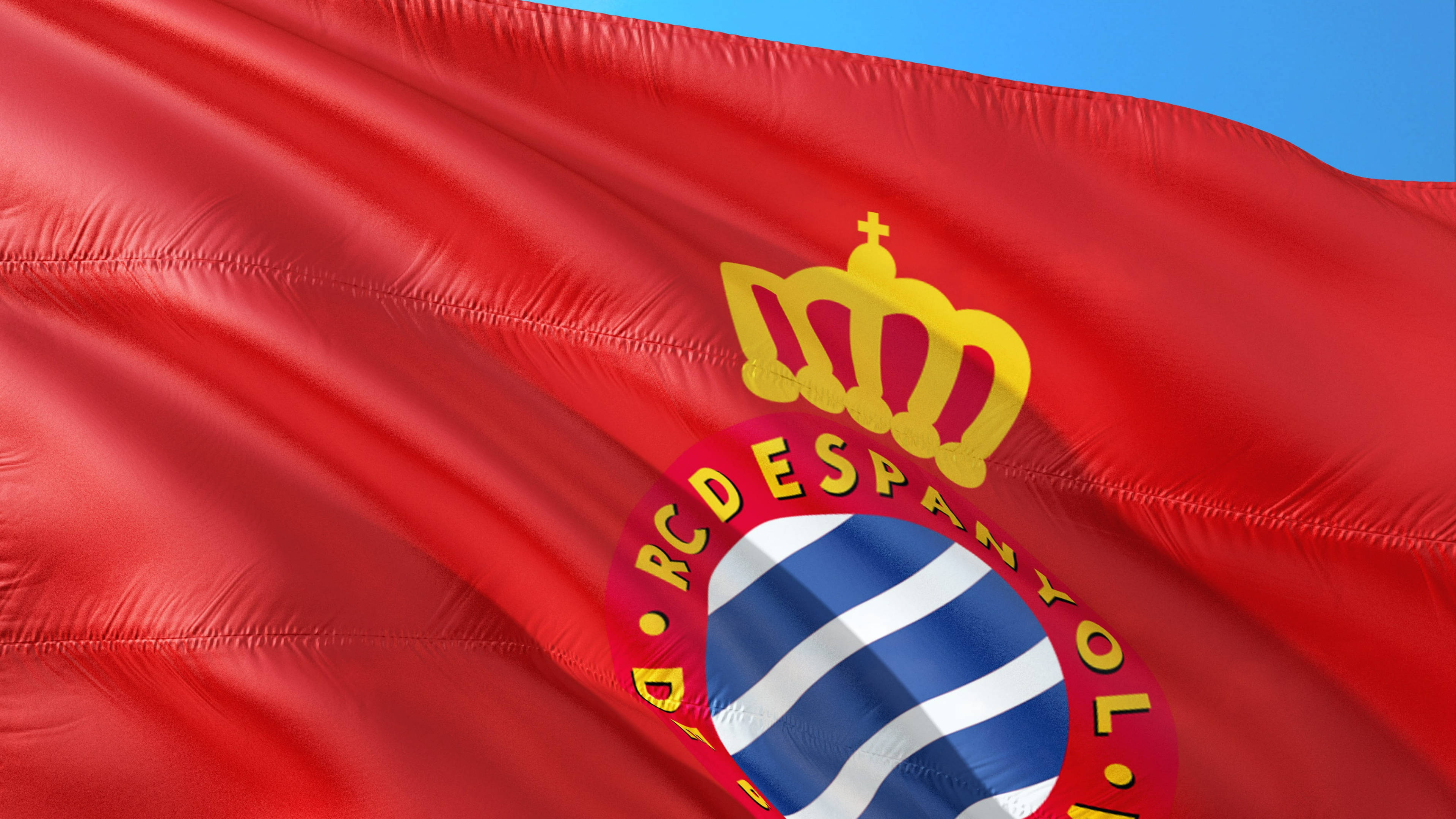 La 桌布 壁紙 — RCD Espanyol 鮮豔的紅旗在風中飄揚，映襯著清澈的藍天，展示帶有皇冠和條紋盾牌的詳細徽章。