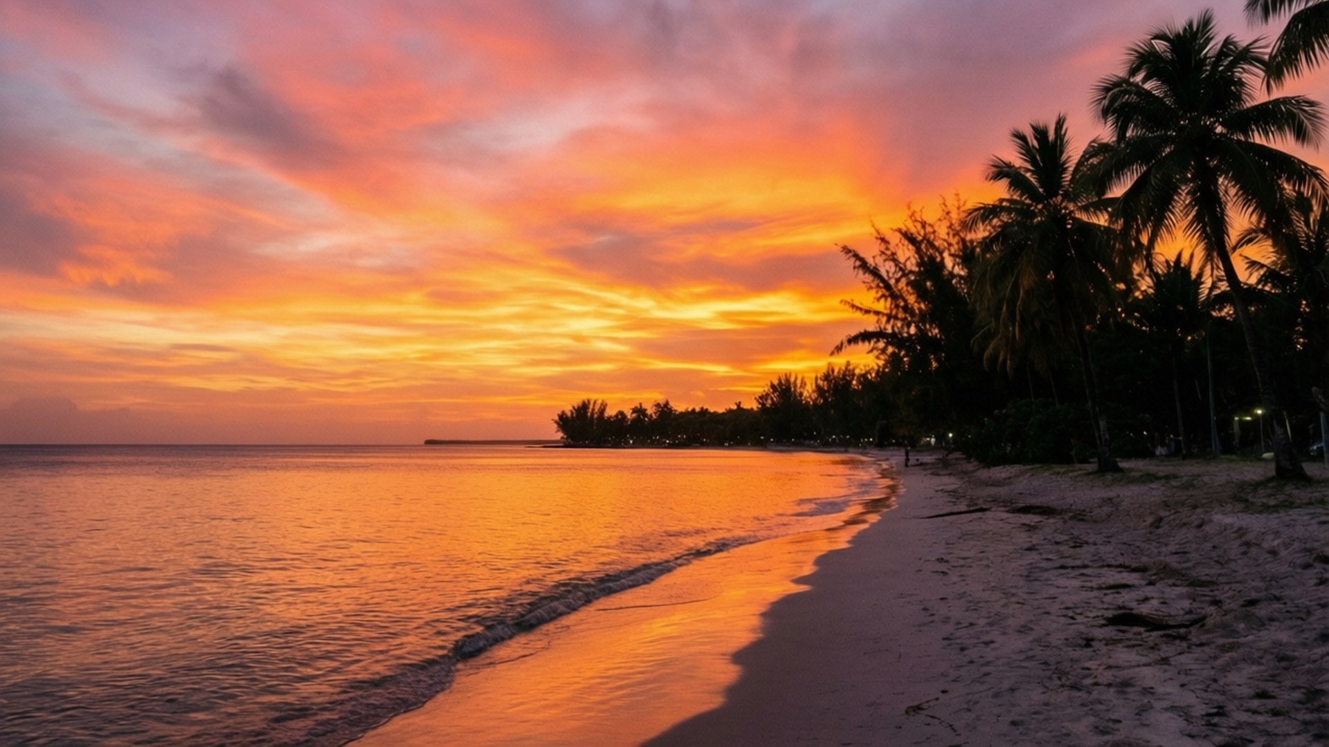 Sunset Serenity at Jamaica 壁紙 — 充滿活力的日落用火熱的橙色和粉紅色調描繪天空，映照在一個寧靜的熱帶海灘上，兩旁是剪影般的棕櫚樹。